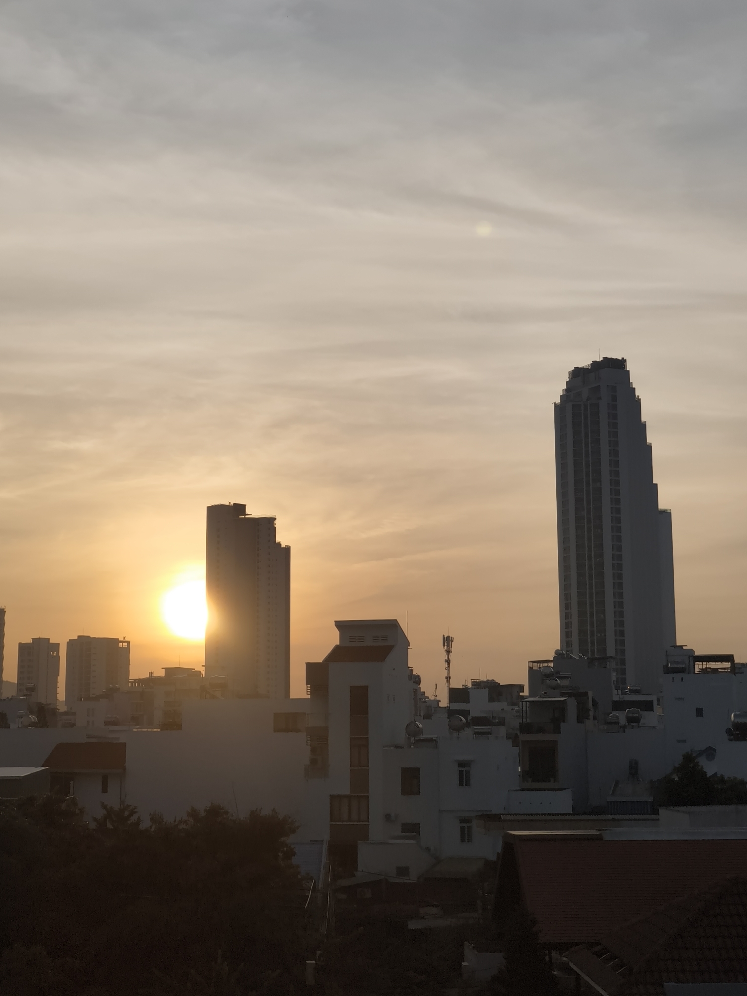 The image depicts a cityscape at sunrise in Bac Nha Trang Ward, Vietnam. The sun, a bright orb, is partially obscured by a tall, slender skyscraper on the right side of the frame, casting a warm, golden light across the sky. Several other buildings of varying heights are visible, some silhouetted against the dawn sky. The architecture suggests a dense urban environment. The sky is filled with soft, diffused clouds that are illuminated by the rising sun, creating a gradient of orange and pale yellow hues. In the foreground, the tops of trees and roofs of lower buildings are visible, suggesting the vantage point is from a higher elevation, possibly a balcony or rooftop. The overall scene conveys a sense of calm and the beginning of a new day in a coastal city.