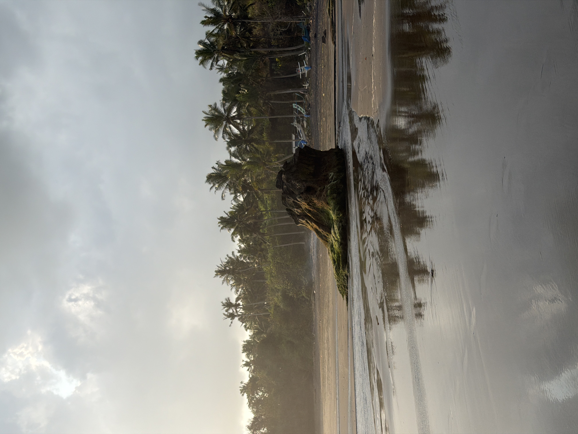 The image depicts a serene beach scene in Selemadeg, Indonesia. The foreground features wet sand reflecting the overcast sky and a line of palm trees in the distance. A large, moss-covered rock sits prominently in the middle ground. Beyond the rock, the beach stretches towards a dense line of palm trees and lush vegetation. Several small, traditional outrigger boats are visible parked on the sand amongst the palm trees. The sky is filled with clouds, with a hint of sunlight breaking through in the upper left portion of the frame. The overall atmosphere is calm and tranquil, characteristic of a tropical coastline.