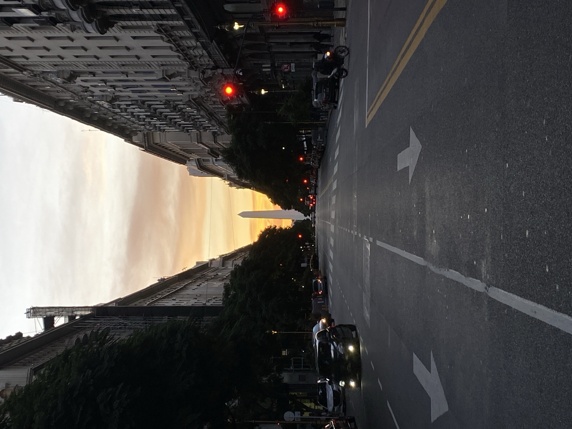 The image shows an avenue in Buenos Aires, Argentina, with the Obelisco monument visible in the distance during a sunset. The avenue is paved and features directional arrows. There are tall buildings on both sides of the avenue, interspersed with trees and streetlights. Several cars and a motorcycle are seen on the avenue, with headlights illuminated. The traffic lights are red.
