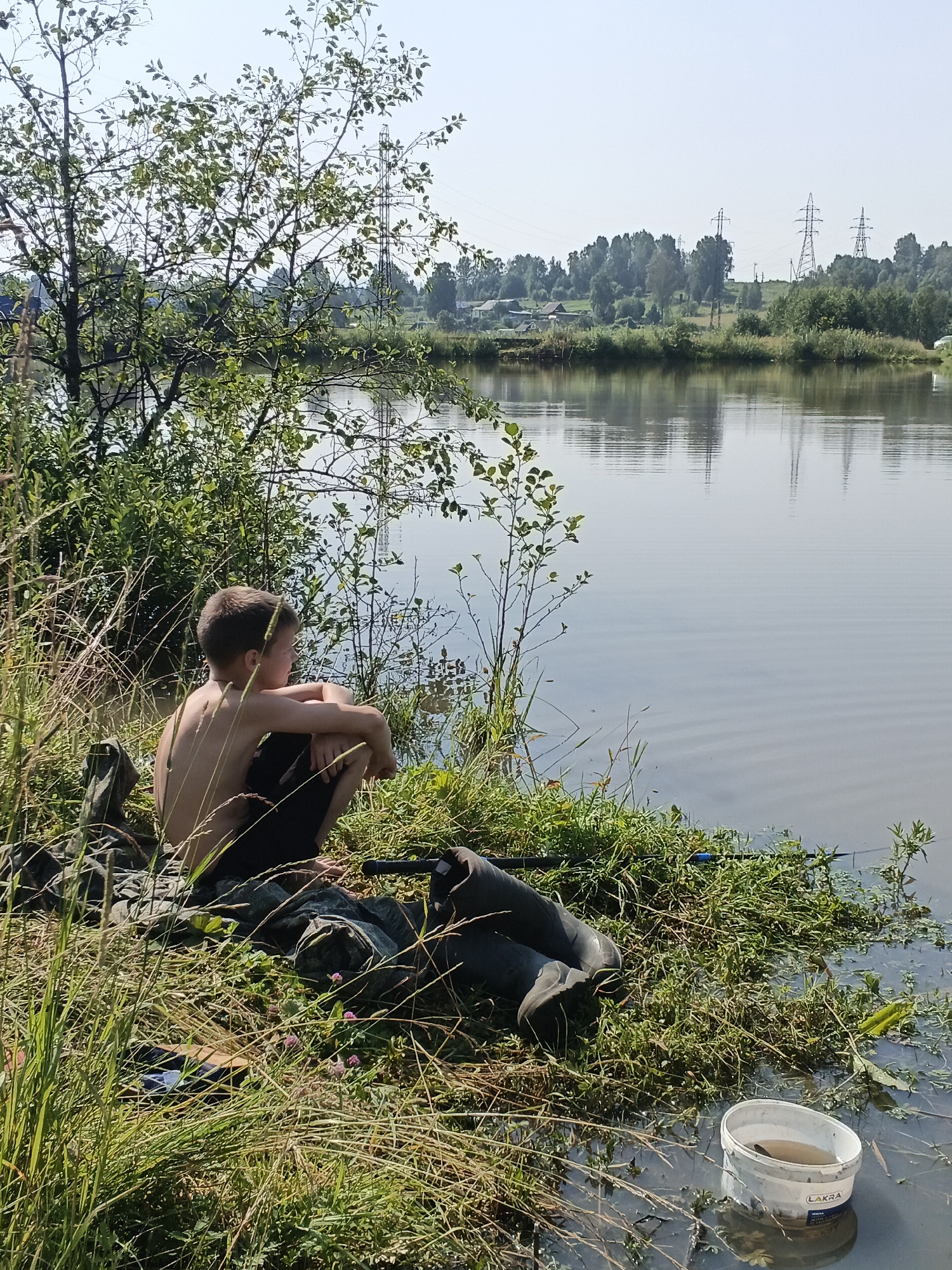 A young boy sits on the grassy bank of a calm body of water, looking out at the reflections on the surface. He is shirtless and wearing dark shorts, with his knees pulled up to his chest. Beside him are fishing gear, including waders and a fishing rod, suggesting he is on a fishing trip. A white bucket, labeled "LAKRA," floats near the edge of the water, containing what appears to be a fish. In the background, across the water, the distant treeline is dotted with power lines and some structures, hinting at a semi-rural or suburban setting. The sky is clear and bright, indicating a sunny day, likely in the late morning or early afternoon given the light. The overall mood is one of peaceful solitude and quiet observation.