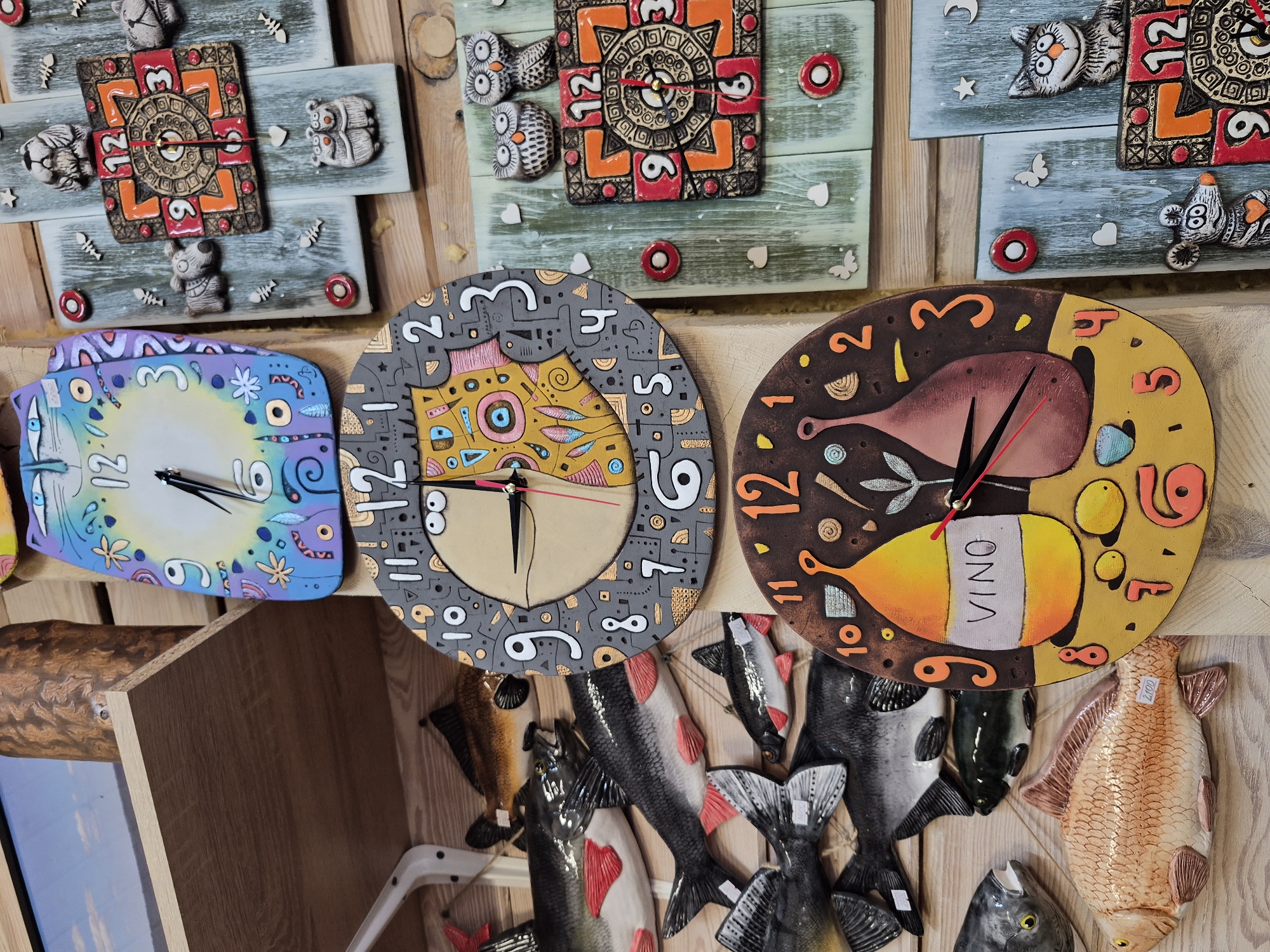 The image displays a close-up view of a collection of decorative ceramic clocks arranged on wooden shelves and a wooden wall. The clocks feature whimsical designs, with some depicting cats, owls, and other animals, as well as abstract patterns and a wine bottle motif. One clock has the word "VINO" written on it. Several ceramic fish sculptures are also visible, hanging on the wooden wall. The overall setting appears to be a shop or market stall, likely selling handmade crafts. The background consists of wooden planks and shelves, with a hint of a blue sky visible on the top left. There are no people present in the image.