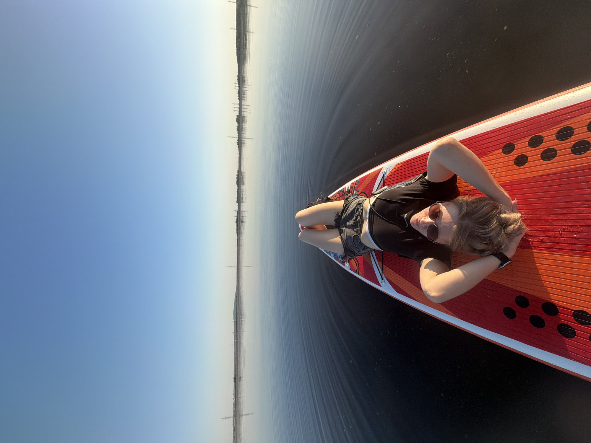 The image depicts a person lying on a paddleboard floating on a body of water. The person is wearing sunglasses, a black top, and shorts. Their arms are positioned behind their head. The paddleboard is orange and red with black polka dots. In the background, a skyline of the city of Chaikovsky, Russia, is visible, characterized by buildings and power lines, suggesting an urban or industrial landscape along the water's edge. The sky is blue and clear. The water appears calm with ripples.