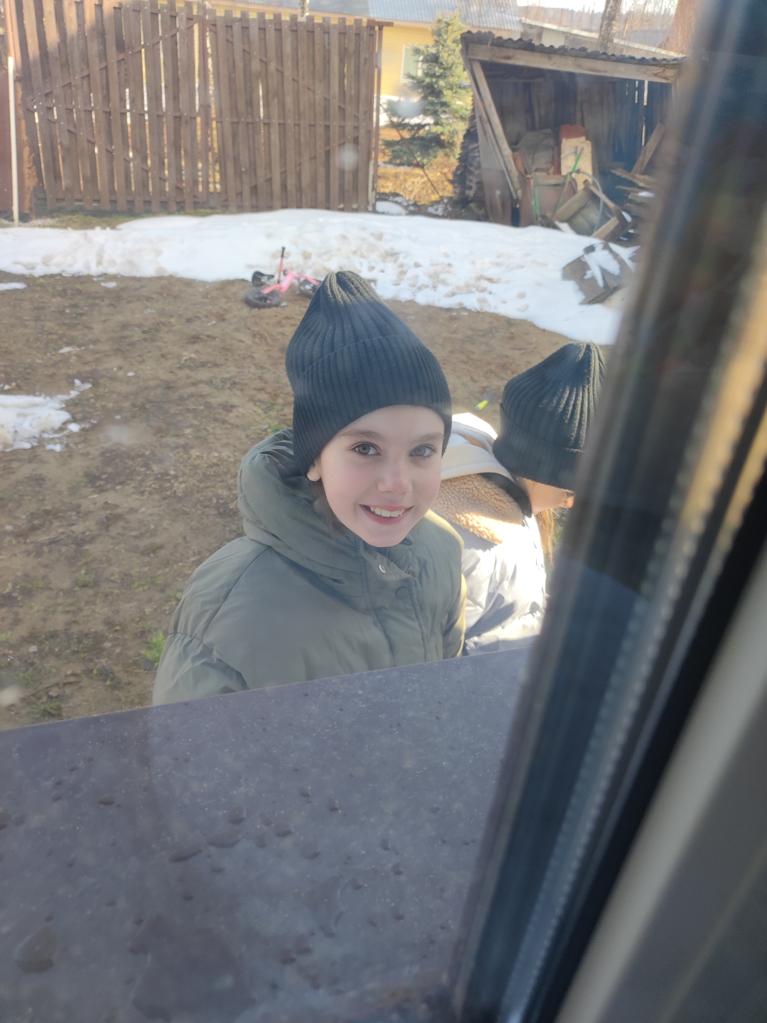 Two children, both wearing dark knit caps, are seen outdoors through a window. The child in the foreground is smiling and wearing a green puffer jacket. The second child is partially visible to the right, wearing a light-colored jacket with a fur-lined hood. The background includes a wooden fence, patches of melting snow, a small children's bicycle, a building or shed, and some greenery. The setting is likely residential, possibly in Votkinsk, Russia.