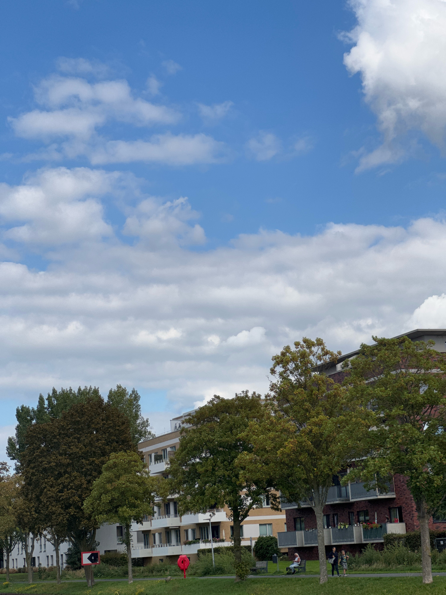 This is a photograph of a residential building complex in Cleves, Germany, viewed from a waterfront area. The sky is bright blue with scattered white cumulus clouds. In the foreground, a grassy area slopes down to the water, which is not visible. Several trees with green foliage line the path in front of the buildings. A red, heart-shaped balloon is situated on the grass. A red and white directional sign with a black arrow pointing left is also visible. A light-colored building with multiple floors and balconies extends across the middle ground, featuring windows, railings, and potted plants. Some people are present on a paved walkway in front of the building; one person is sitting on a bench, and two others are standing. The buildings are modern in design, with a mix of light-colored facades and red brick sections.