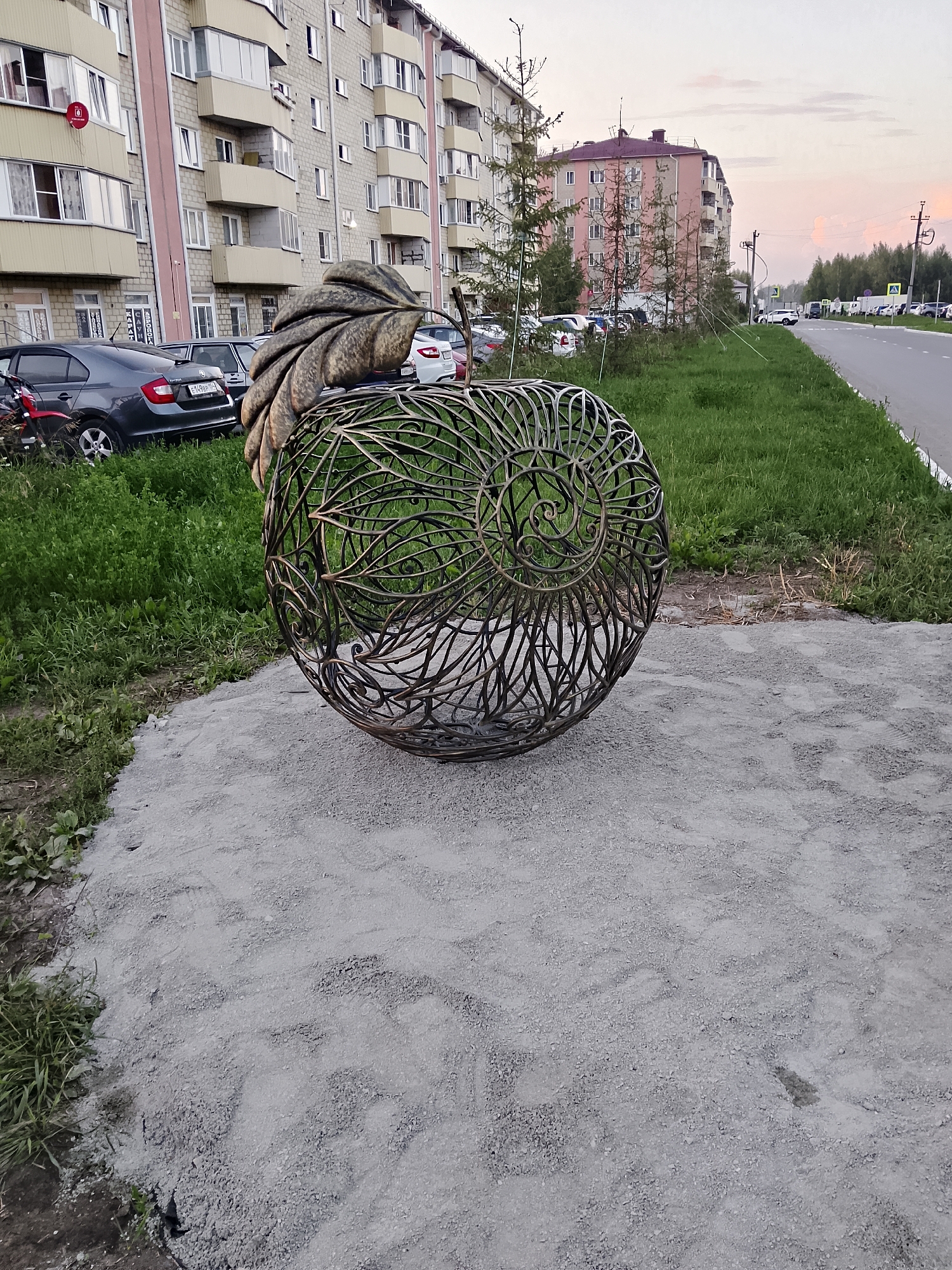 A decorative metal sculpture resembling an abstract fruit, possibly an apple or pear, with a large leaf on top, is situated on a patch of sandy gravel in front of a multi-story apartment building in Oktyabrskiy, Russia. The sculpture is made of dark metal rods twisted into a spherical shape, with leaf-like patterns visible within its structure. Beyond the sculpture, a grassy area with some young trees separates it from a paved road. Several parked cars are visible on the left side of the image, in front of the building, and along the right side of the road, which stretches into the distance. The sky above is a soft, dusky hue, suggesting either early morning or late evening light. The apartment building is pale yellow with light pink accents and has numerous windows and balconies. The overall scene depicts an urban residential area with public art.
