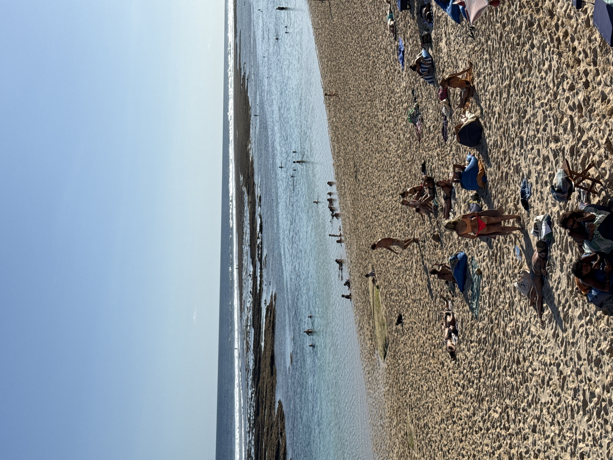 This image captures a lively, sunny beach scene in Kuta Selatan, Indonesia, viewed from a slightly elevated perspective looking down across the shoreline and ocean. The scene is dominated by a wide stretch of light brown, textured sand on the right, and the calm, clear blue-green ocean on the left, meeting a bright, clear blue sky at the horizon.
Numerous people are scattered across the sandy beach, primarily engaged in sunbathing and relaxation. Many are lying down on towels or directly on the sand, dressed in swimwear. Small beach umbrellas, towels in various colors (blue, white, patterned), and personal bags are visible around them. The sand shows distinct ripple patterns, indicating the movement of water or wind.
In the shallow, translucent water along the shore, several individuals are wading or standing, enjoying the ocean. Darker patches visible within the water suggest rocky areas or reef formations. Further out, small waves can be seen.
The weather is bright and clear, with abundant sunshine indicated by the deep blue sky and the distinct shadows cast by people and objects on the sand, suggesting it is likely mid-day. The overall atmosphere conveyed is one of leisure and enjoyment. No visible text is present in the image.