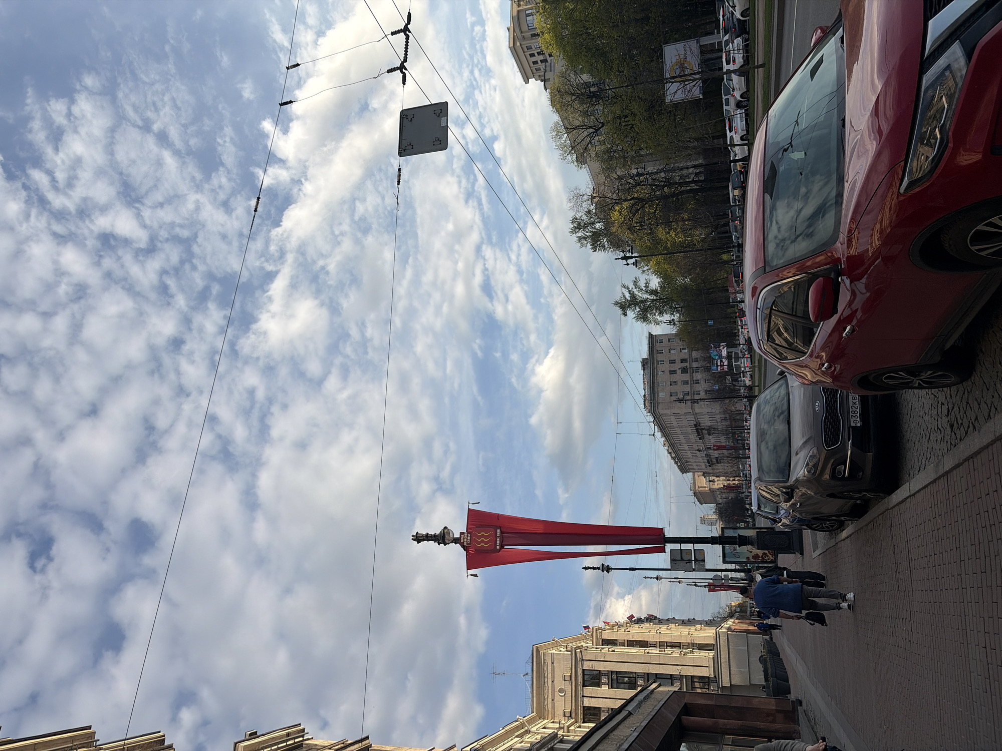 The image is an eye-level street scene in Saint Petersburg, Russia. In the right foreground, there's a red car parked along the edge of a brick sidewalk. Further down the sidewalk, people are walking. Cars are parked along the street beyond the sidewalk, and buildings line the street. On the left of the image, there are overhead electrical wires. A red banner with a coat of arms on it is suspended between two lampposts. The sky is filled with puffy white clouds against a blue sky.
