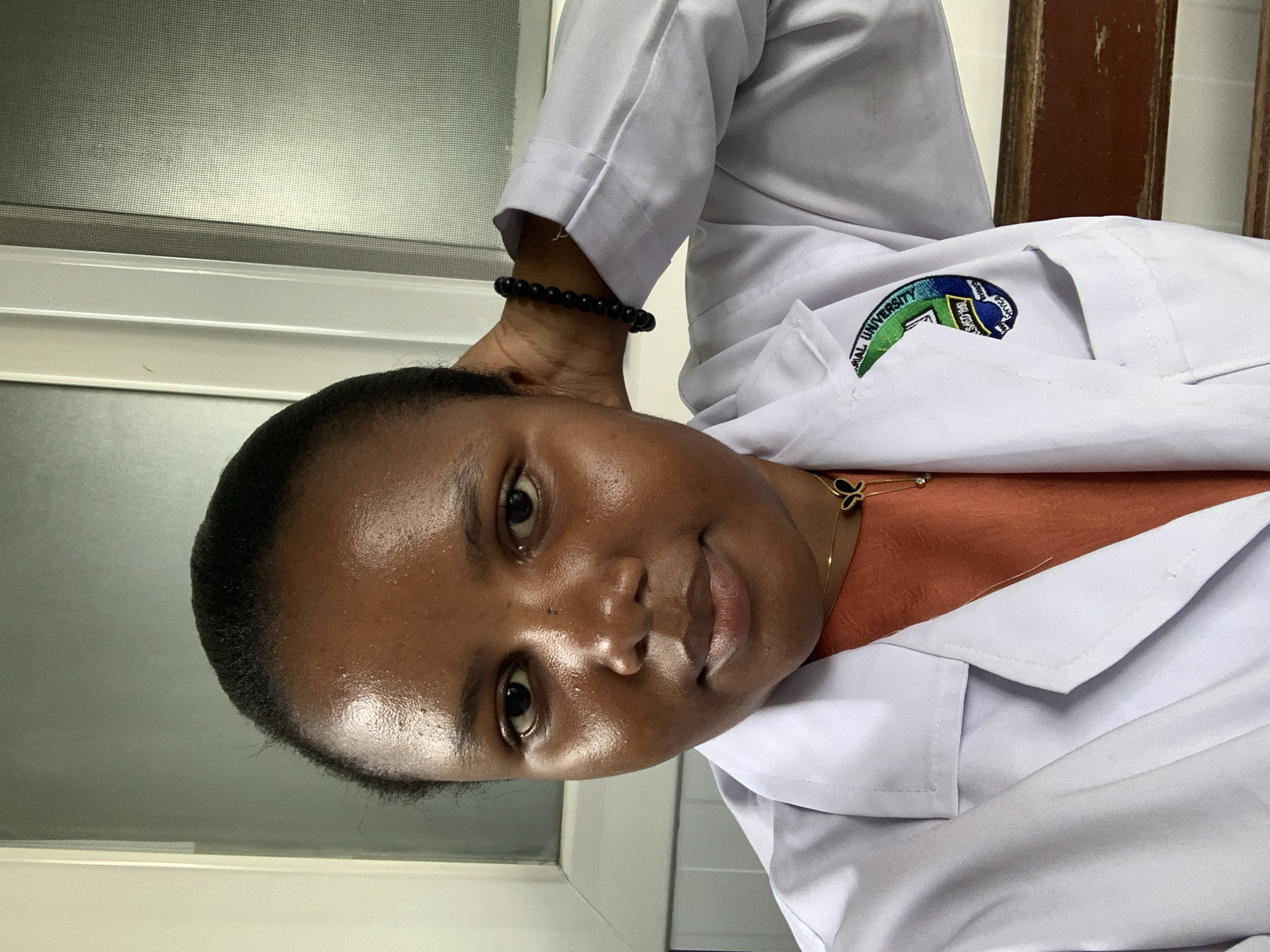A young adult female of African descent is depicted in a medium close-up, indoors, in what appears to be a university or medical facility setting within Dar es-Salaam, Tanzania. She is looking directly forward, maintaining a neutral expression. Her dark hair is pulled back, and her skin appears somewhat lustrous.
She is wearing a white lab coat over an orange-brown top. A gold-colored chain necklace with a black and gold butterfly pendant is visible around her neck. On her left wrist, she wears a bracelet composed of dark, spherical beads. Her left hand is positioned with fingers touching the back of her head or neck area.
The white lab coat features an embroidered circular emblem on the left side of the chest (viewer's right). The emblem includes green, yellow, and blue sections, with text reading "NATIONAL UNIVERSITY" at the top, a possible "ESTABLISHED 1997" in a yellow banner, and "RESEARCH AND SERVICES" on a blue arc at the bottom.
The background consists of a white-framed translucent glass panel or window to the subject's left. To her right, a dark brown, horizontally oriented element, possibly the top rail of a wooden bench or chair, is visible. The immediate wall surface is light-colored.