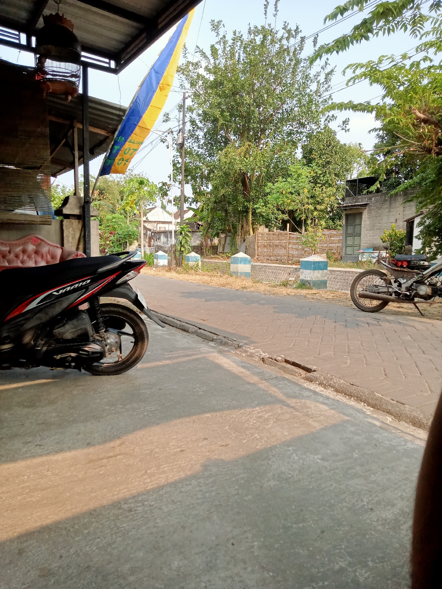 In the city of Majan, Indonesia, a scene unfolds on a street bathed in the warm glow of late afternoon sun. The foreground is dominated by a concrete walkway, with a dappled shadow stretching across it, hinting at the low angle of the sun. To the left, a red and black motorcycle is parked, its sleek design contrasting with the rustic surroundings. Beyond the motorcycle, a portion of a pink upholstered bench is visible, suggesting a seating area.
The street itself is paved with interlocking bricks, with a raised concrete curb separating it from the walkway. Along the street, two cylindrical bollards, painted in alternating bands of white and blue, mark a boundary. In the background, a silver motorcycle is parked further down the street.
The environment is characterized by lush greenery, with a variety of trees and plants creating a natural backdrop. A weathered wooden fence with woven bamboo panels lines the street in the mid-ground. A small building with a grey brick facade and a green-framed window stands on the right. Overhead, a metal-framed awning is visible, with a birdcage suspended from it. A blue and yellow flag, with some text partially obscured, hangs limply, possibly from a nearby pole. Utility poles and wires crisscross the sky, indicating the presence of infrastructure. The overall impression is one of a quiet, residential street in a tropical climate, with a blend of man-made structures and natural elements.
