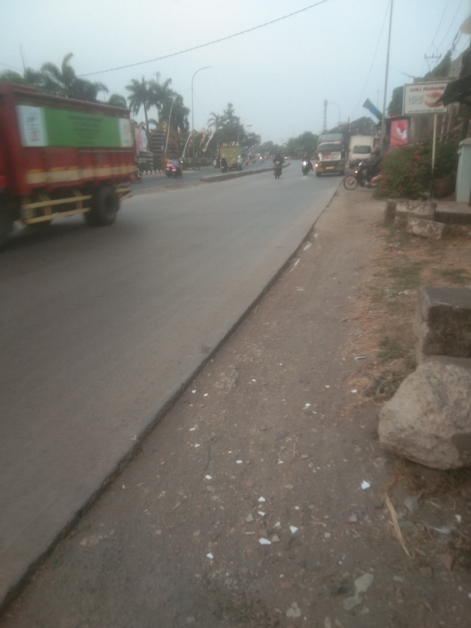 The image depicts a street scene in Subang, Indonesia, likely during daytime or early evening given the ambient light. A red truck with a green tarp-covered cargo is on the left side of the road, moving away from the camera. The road itself appears to be asphalt, with a dirt and gravel shoulder on the right. Several motorcycles are visible on the road, some with riders. In the background, there are palm trees and other foliage. A building with signage, including what appears to be an advertisement for dental services, is situated on the right side of the road. The sky is overcast, contributing to a diffused light. Several large rocks are present on the shoulder of the road, along with scattered debris. The overall impression is that of a typical urban or semi-urban environment in Indonesia.