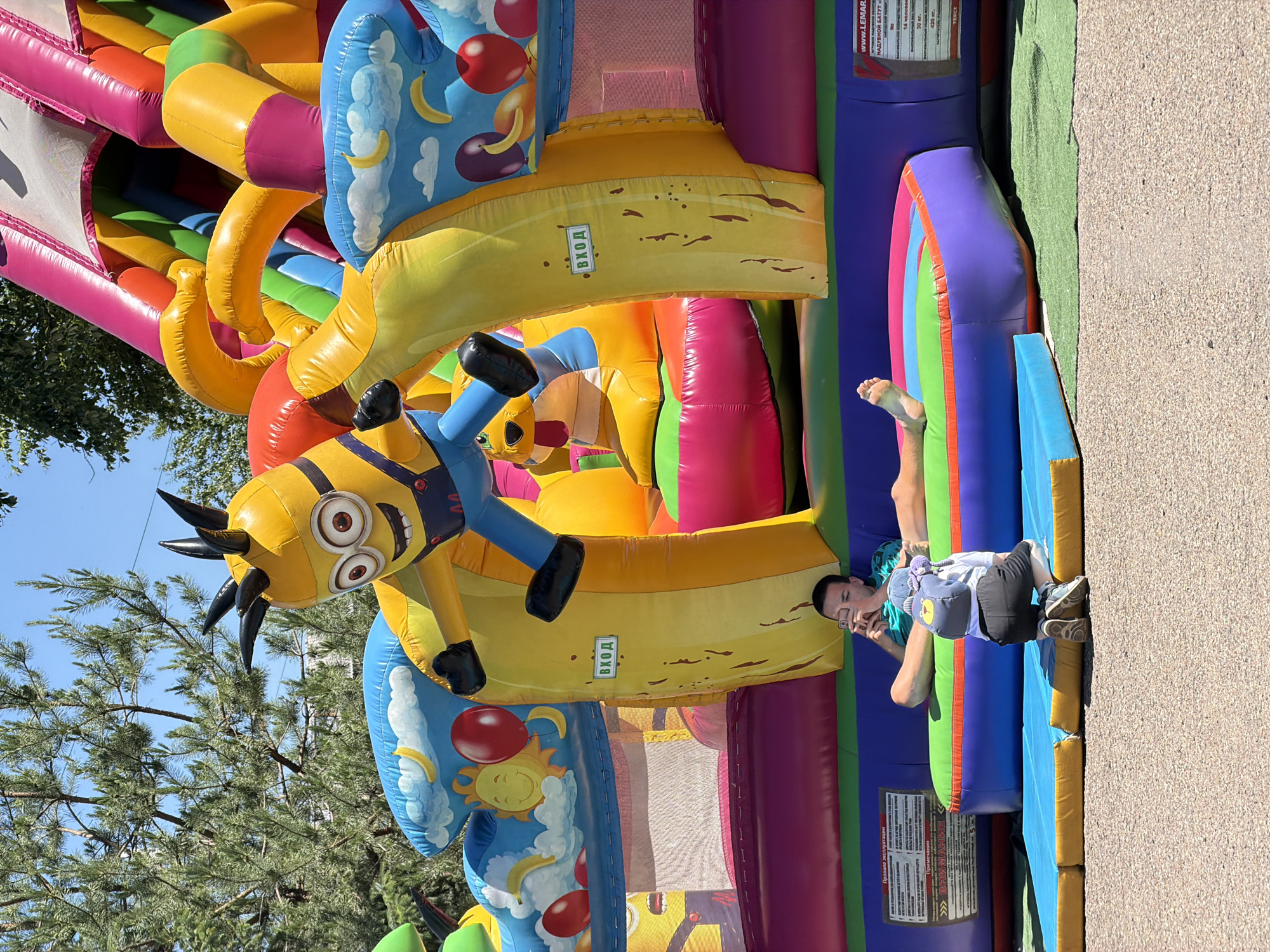 In Birobidzhan, Russia, on a sunny day, a large, colorful inflatable playground is set up outdoors. The inflatable structure features a giant Minion character from the Despicable Me franchise at its entrance. In the foreground, a young child wearing a white t-shirt and black shorts is kneeling, facing away from the camera, holding a smartphone and appearing to take a picture. Behind the child, a young man is sitting with his legs extended, looking at his own smartphone. The inflatable structure itself is a complex arrangement of slides and bouncy areas, adorned with cartoon images of the sun, moons, and fruit. The ground around the inflatable is a mixture of gravel and a green mat.