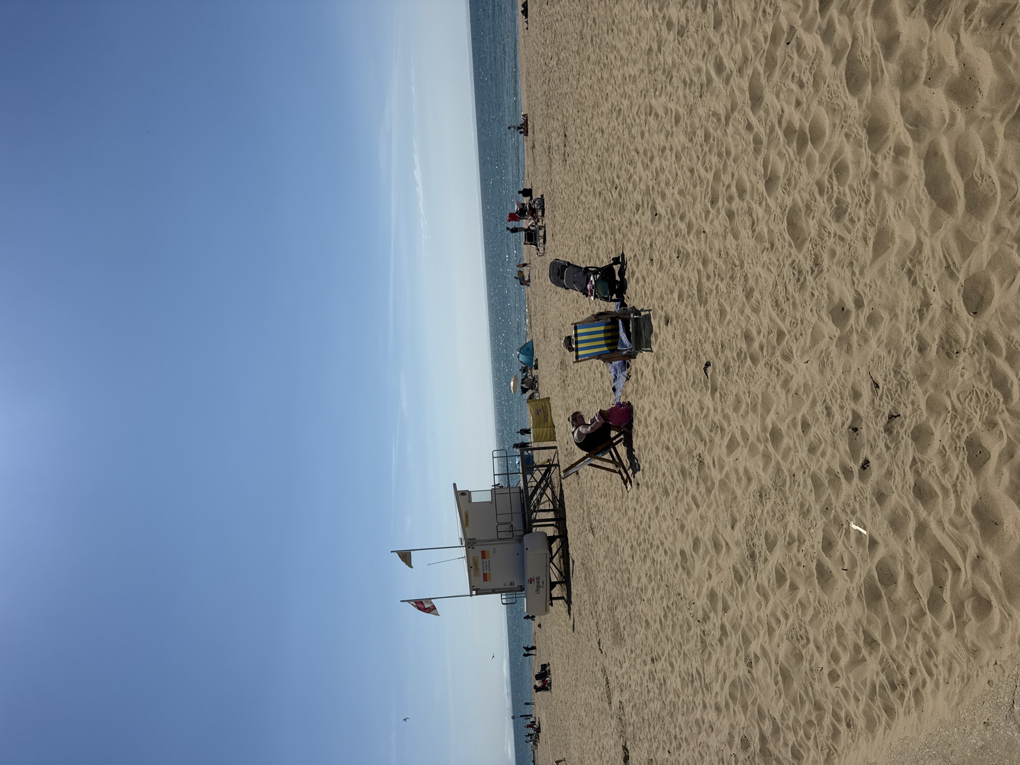 This is a vertical, eye-level shot of a sandy beach with the ocean in the background. The sky is clear and blue, suggesting a sunny day. The time of day appears to be midday or early afternoon, given the bright sunlight casting shadows. The location is Bournemouth, UK, indicated by the presence of a lifeguard tower with text that reads "Lifeguards" and the Union Jack flag.
The beach is populated with several people. In the foreground, a person is relaxing in a beach chair with a yellow and blue striped umbrella behind them. Further back, there are several other individuals and groups scattered across the sand, some sitting in chairs, others standing. A stroller is parked on the sand. The lifeguard tower is a prominent feature, situated on the beach near the water's edge. Two flags are attached to the tower. The sand is textured with footprints and ripples, evidence of recent activity. The ocean is calm and reflects the sunlight. The overall atmosphere seems relaxed and leisurely, characteristic of a typical beach day.