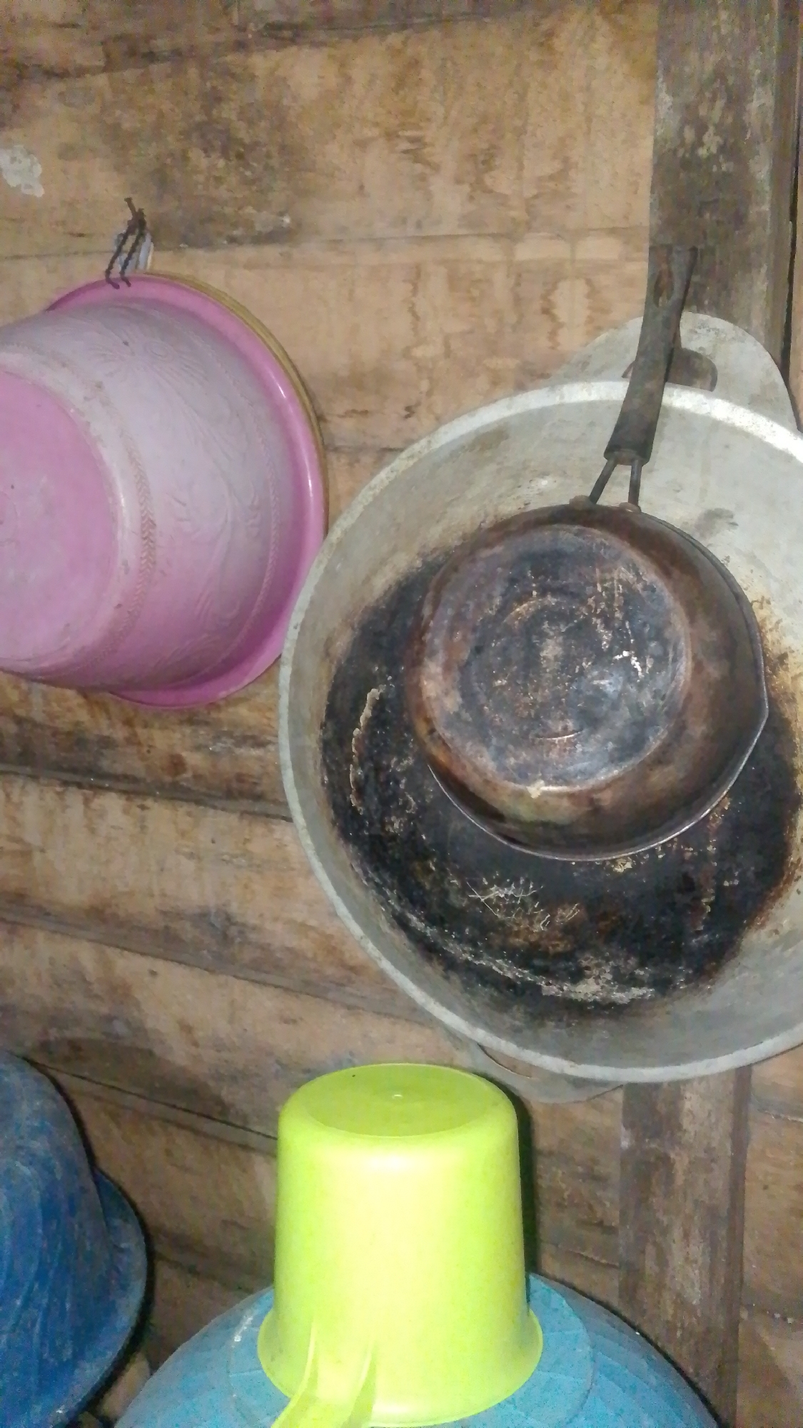 An indoor scene, likely a kitchen, in Olak-Olak Kubu, Indonesia, features several household items against a wooden wall. A pink plastic basin hangs on the left. To its right, a large, tarnished metal basin holds a blackened frying pan, its handle propped up. In the foreground, a bright green plastic mug sits atop a blue container. The environment appears rustic, with visible wood grain and staining on the walls.
