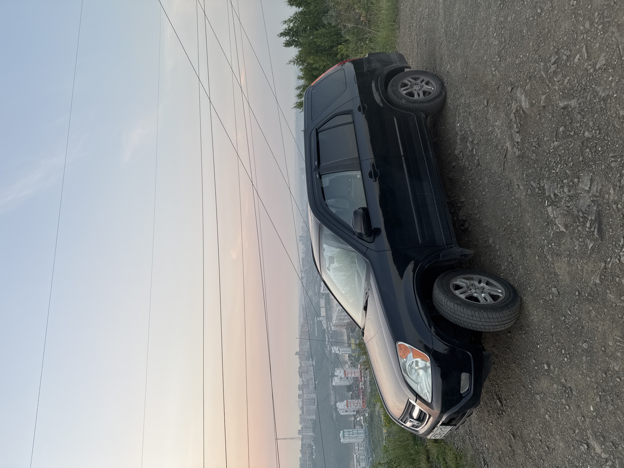 The image shows a black Honda CR-V parked on a dirt road with a city and power lines in the background. The car is angled, with its front facing towards the right of the frame. The sky is a soft gradient of pale blue and orange, suggesting either sunrise or sunset. Several thick power lines run diagonally across the sky and foreground, creating a graphic element. In the distance, a sprawling city with numerous buildings is visible, partially obscured by a hazy atmosphere. The setting appears to be a hillside overlooking the city, with some greenery and trees present. The ground beneath the car is composed of loose gravel and dirt. There is no visible text in the image. The overall mood is serene and quiet, with the car appearing to be stationary.