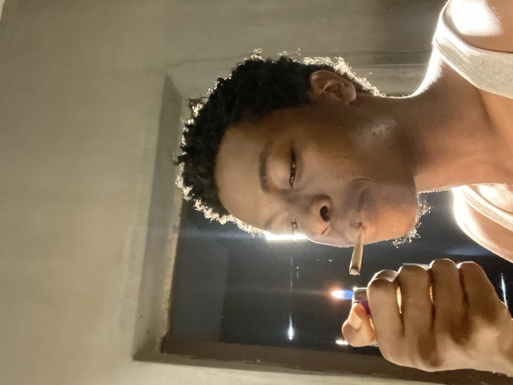 A person with dark skin and short, curly hair is shown in profile, lighting a cigarette with a purple lighter. The background is dimly lit and appears to be an indoor setting, possibly a doorway, with a bright light source visible behind the person. The person is wearing a white tank top. The image is taken at eye level, focusing on the person's face and hand holding the lighter and cigarette. The scene suggests a casual or private moment. The location is identified as Nkpor, Nigeria.