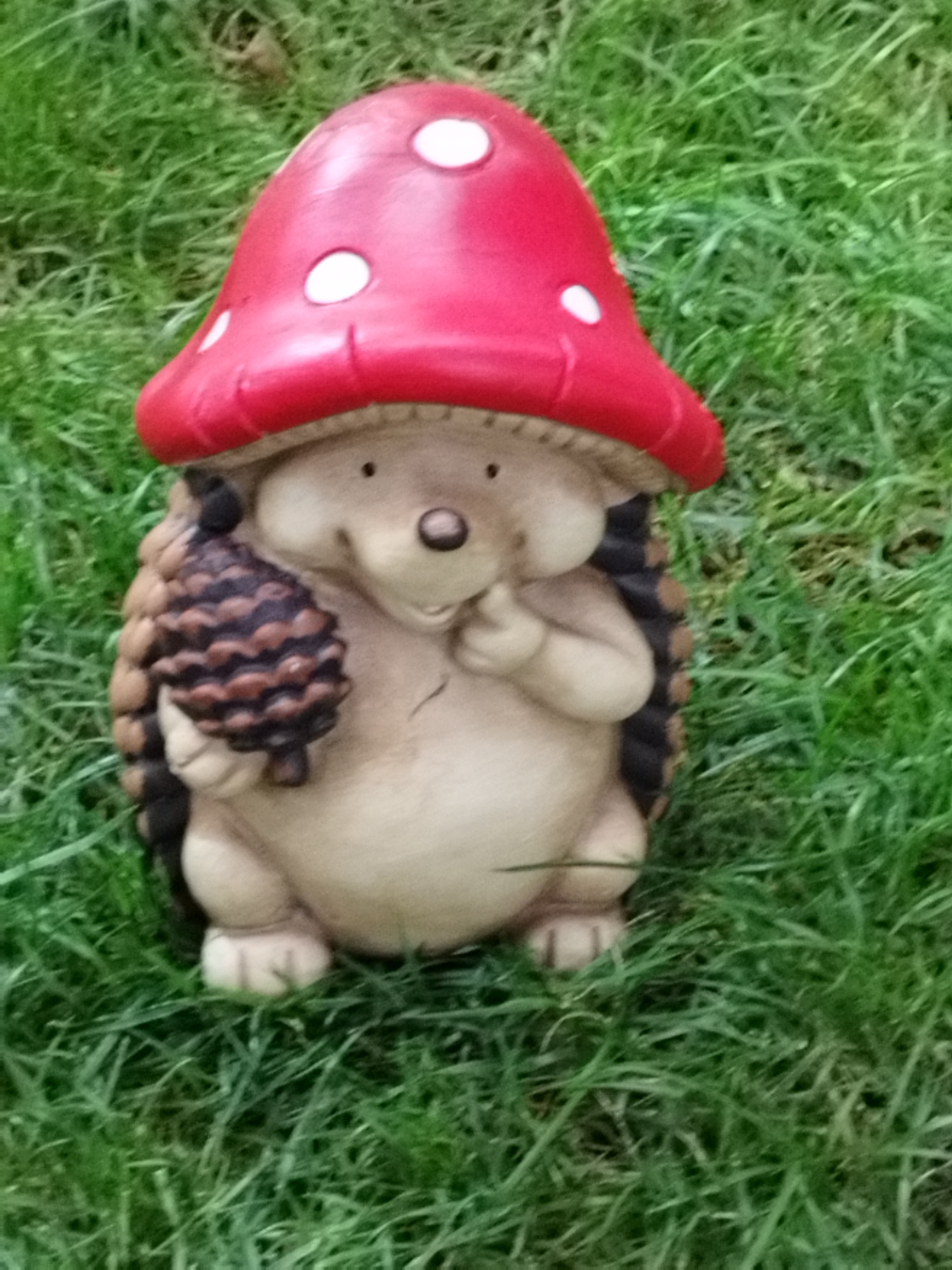 An outdoor, close-up, eye-level, slightly tilted shot features a garden gnome figurine in the shape of a hedgehog wearing a red mushroom cap. The gnome is made of ceramic and has a light brown body with darker brown quills. It is holding a brown pine cone in its right hand and has its left index finger to its mouth. The figurine is placed on bright green grass. The background is entirely out of focus, showing only the green blades of grass. No people or animals are present. The location is not specified within Bucharest, Romania.