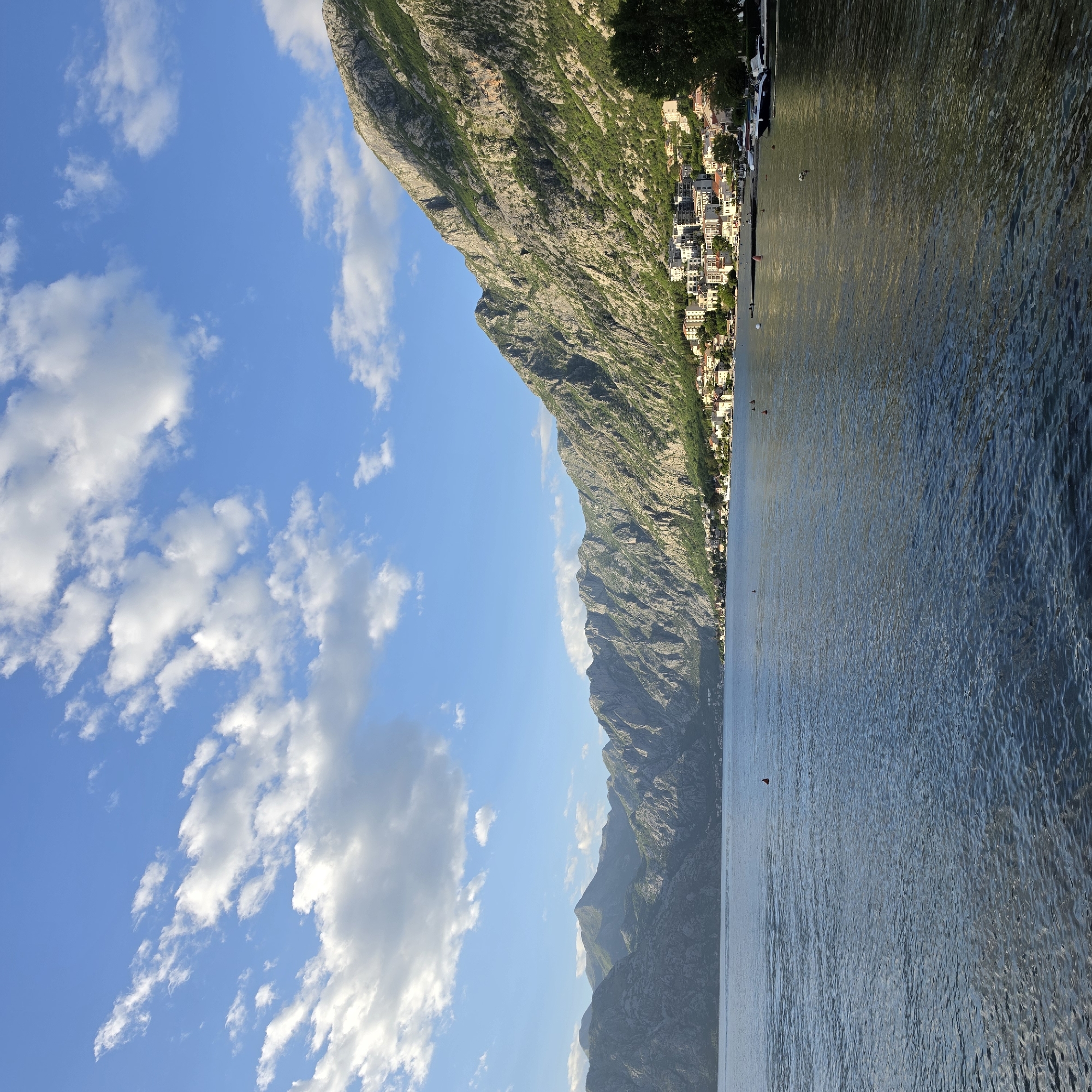 The image depicts the landscape of Dobrota, Montenegro. The photograph, taken outdoors during daylight, prominently features a large, rugged mountain range on the left side, partially covered with vegetation and exhibiting rocky terrain. Adjacent to the mountains, a dense collection of buildings and houses aligns along the coastline, suggesting a built-up urban area or town. A vast body of water fills the right side of the image, with gentle ripples and waves on the surface. The sky occupies a significant portion of the photograph, with a bright blue hue and scattered white clouds. The overall scene conveys a natural landscape juxtaposed with the human development of the town.