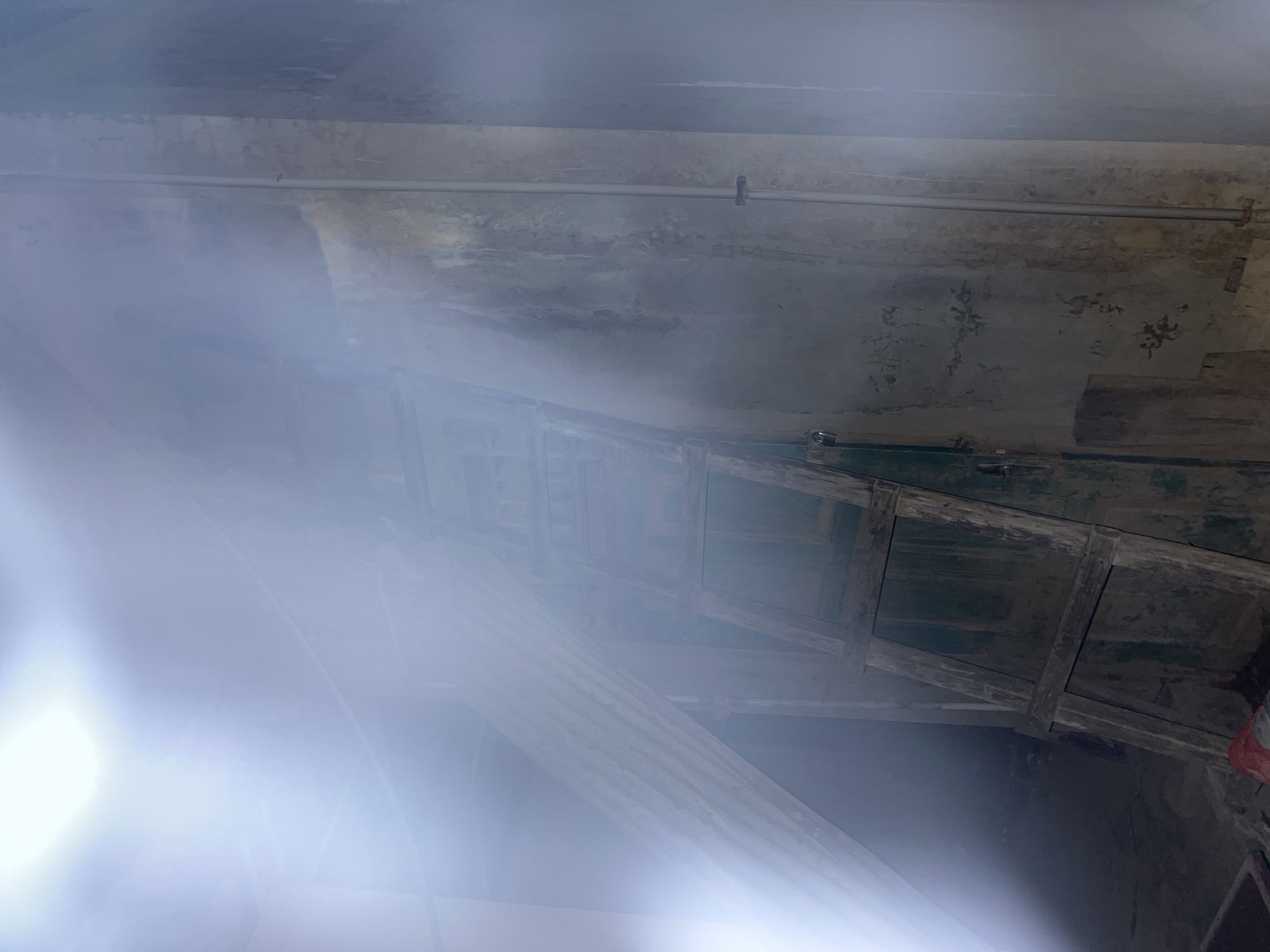 This is an interior, low-angle shot of what appears to be a dimly lit, possibly abandoned or under-construction space. The primary subjects are architectural elements. We see a rough, distressed concrete ceiling with visible cracks and peeling paint, suggesting age or disrepair. A metal pipe runs horizontally across the ceiling, attached by clips.
Below the ceiling, a wooden structure resembling a dilapidated staircase or walkway is visible. It has horizontal wooden planks supported by vertical beams. The wood is weathered and appears to be painted in a dark, possibly green, color in some sections. There are also vertical wooden supports and what look like wooden slats forming a lattice or railing.
A bright, hazy light source is prominently in the lower left of the frame, causing significant lens flare and obscuring details in that area. This light creates a diffused, ethereal atmosphere. The overall impression is one of neglect and decay, with a sense of mystery due to the obscured areas. There is no visible text, people, or clear indication of the time of day or weather outside. The setting evokes a feeling of being in a forgotten or utilitarian space, perhaps a disused factory, warehouse, or basement.