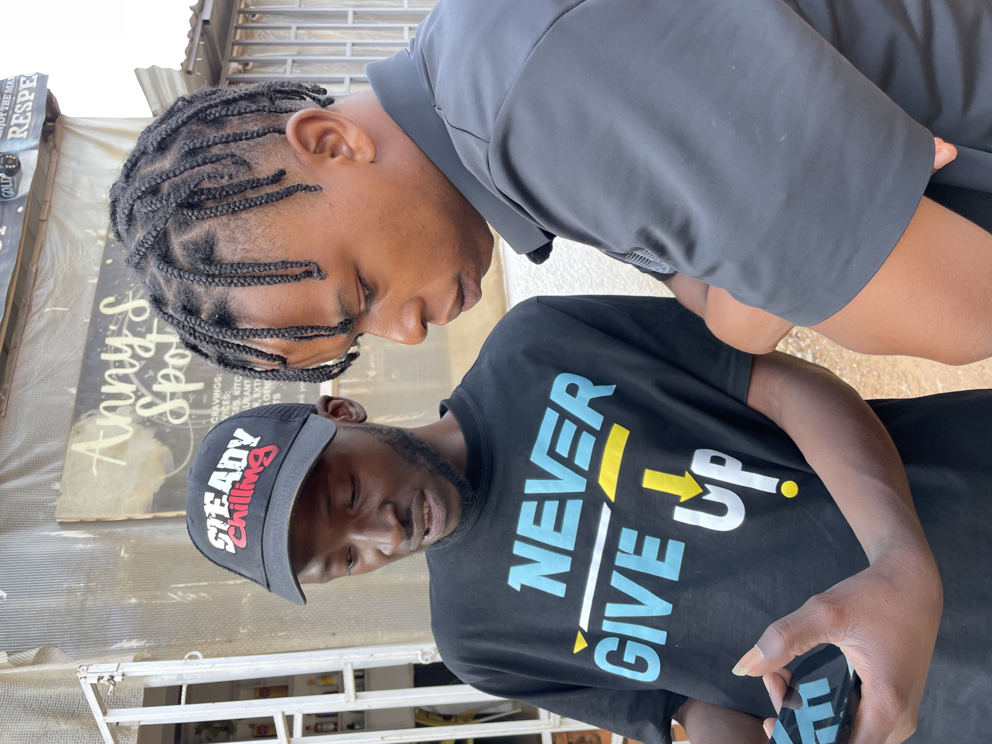 Two men are depicted in a close-up, vertical shot, appearing to be engaged in a shared activity in an outdoor setting in Barakin Akawo, Nigeria. The scene is brightly lit, indicating daytime.
The man on the left, positioned higher in the frame, has dark skin and his hair styled in neat braids (cornrows). He is wearing a dark, possibly black, short-sleeved collared shirt. He is leaning forward, looking intently downwards and to his right, towards the second man.
The man on the right, positioned lower, also has dark skin and is wearing a black short-sleeved t-shirt with prominent text that reads "NEVER GIVE UP" in light blue and yellow lettering. He has a beard and is wearing a black baseball cap with "STEADY Chilling" in white and red text across the front. He is holding a black smartphone in his left hand and is also looking downwards, seemingly at the phone or what the other man is observing.
In the background, behind the men, parts of a storefront or stall are visible. A large, weathered sign features the partial text "Anny S Spot", "CRAVINGS:", "VICES:", "TES, KITC", "URANT", "REXT", "ENJOY THE MAN", and "RESPE". A small "GOLD" sign is also visible. The structures behind them include metal grating and possibly corrugated roofing, typical of an urban or semi-urban outdoor commercial area.