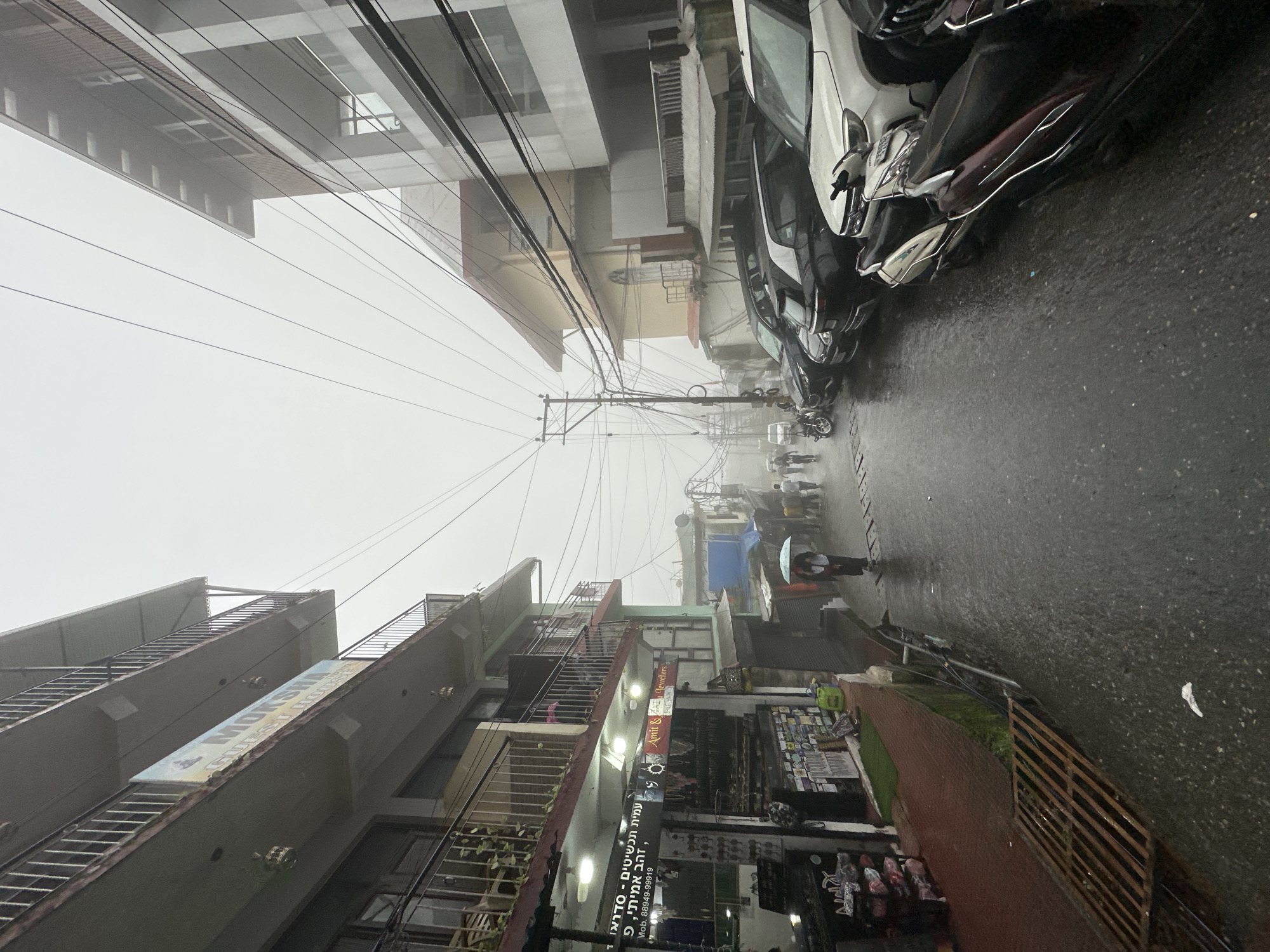 The image depicts a narrow, wet street in a foggy, urban environment. Buildings line both sides of the street, with shops visible on the left and a multi-story structure on the right. The street is paved and wet, indicating rain, with several cars and scooters parked along the right side. A few people are walking in the street, some with umbrellas. The overall atmosphere is one of a cloudy, possibly rainy day in a commercial area. The sign on the left building says "MOKSHAM GUEST HOUSE" and "Amit & Amit Jewellers". There are also signs in Hebrew.