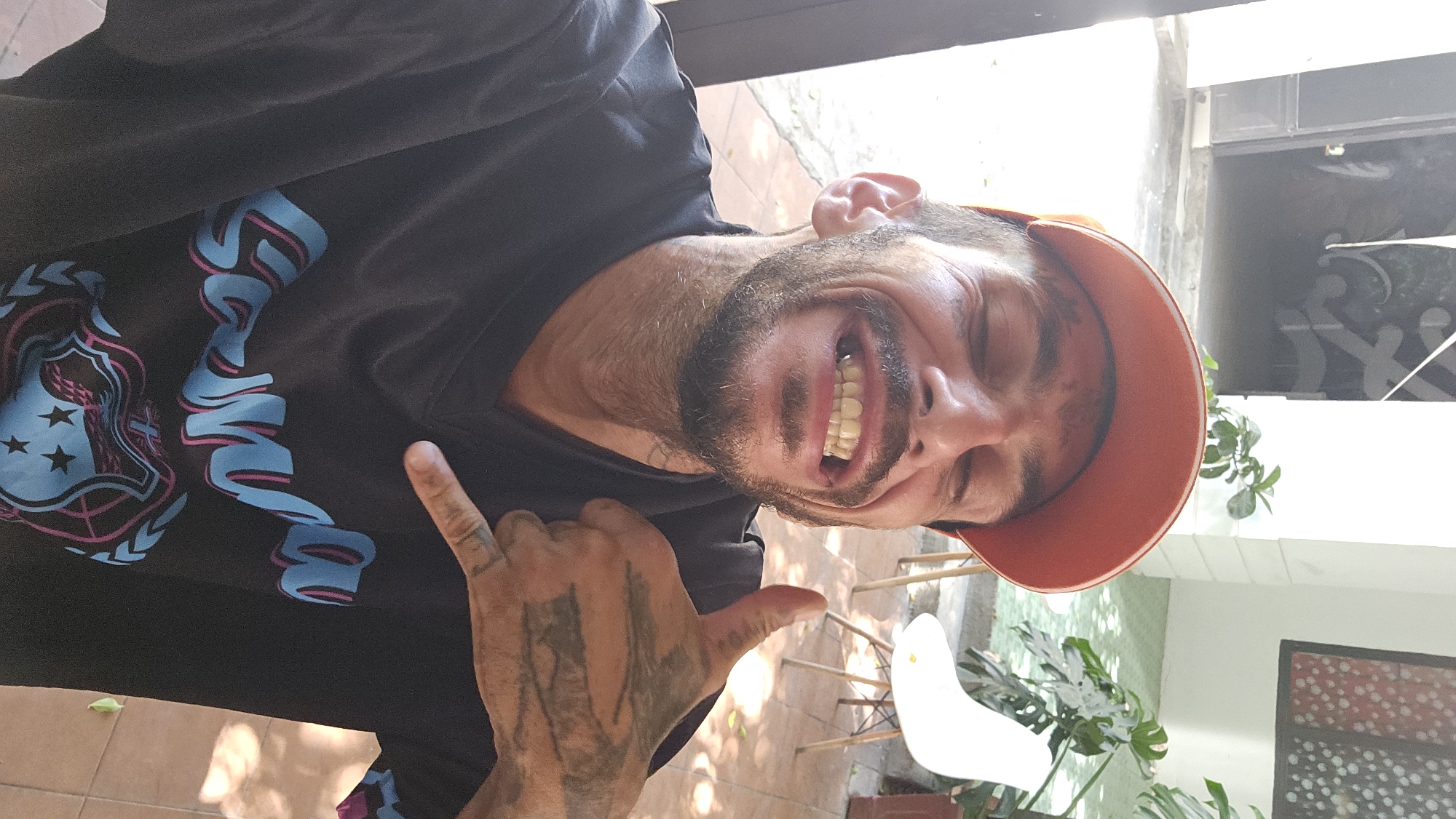 A middle-aged male, wearing a black t-shirt with blue and pink graphics and an orange baseball cap, is smiling broadly with visible dental discoloration. His right hand, heavily tattooed, is making a "hang loose" gesture. He has facial hair. The background suggests an outdoor patio or courtyard, with tiled flooring, a white chair, and potted plants visible. The presence of patterned glass and architectural details suggests a residential or commercial building. There is no clear indication of the location being Nha Trang, Vietnam.