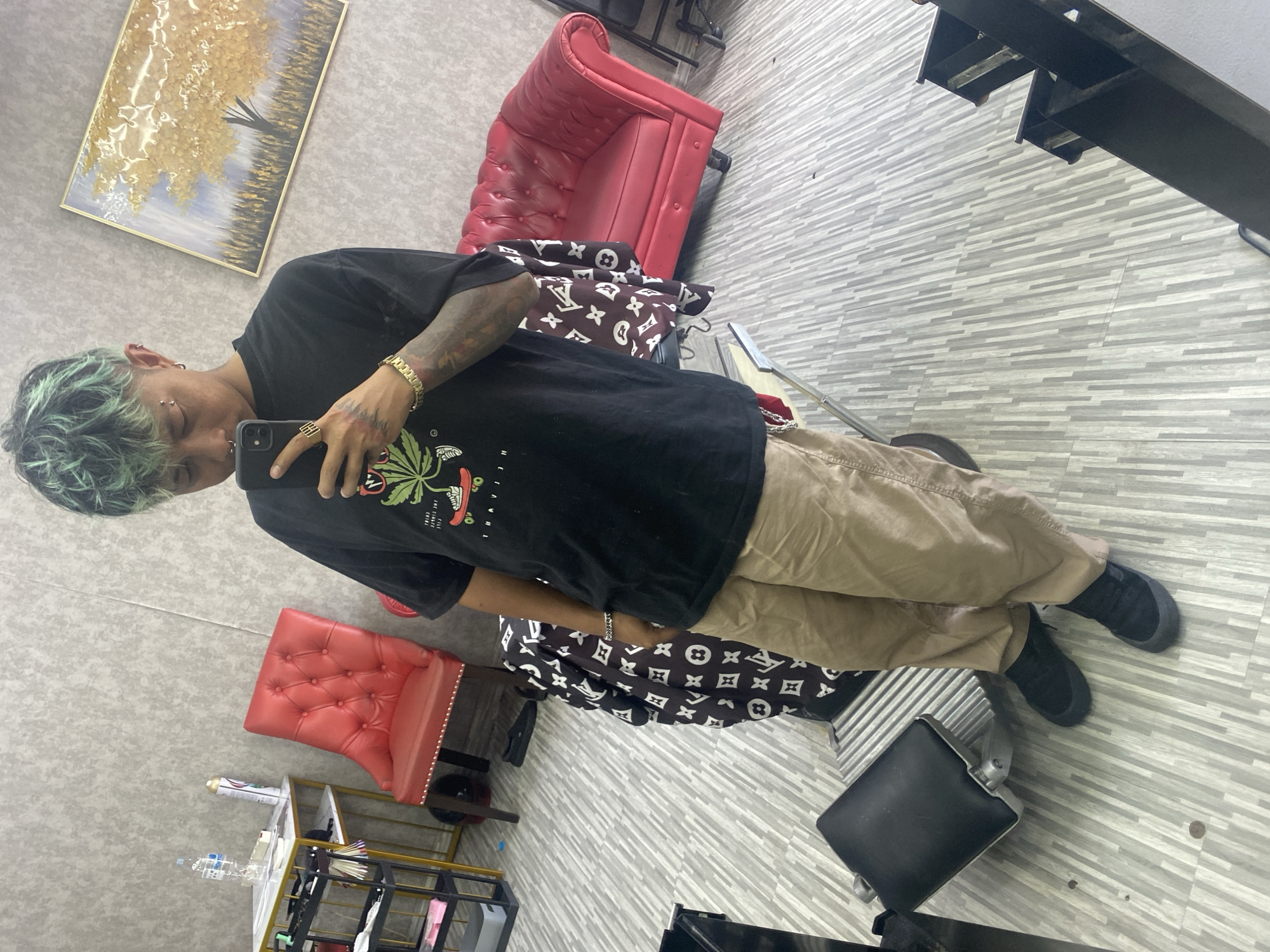 A man is posing for a photo in a brightly lit indoor space. He is facing the camera at an angle, holding a smartphone in his right hand and appearing to take a selfie. He has green-tinted hair, tan skin, and multiple piercings in his ears and eyebrows. He is wearing a black t-shirt with a graphic print, tan pants, and black sneakers. His left arm is tattooed, and he is wearing a gold bracelet on his right wrist and a silver bracelet on his left wrist.
In the background, there is a red, button-tufted chair, a decorative LV-printed blanket, and a painting on the wall. A black metal rack and other salon-style furniture are also visible. The floor is covered in light gray and white striped tiles. The image is taken at a slight angle and appears to be in a salon or similar establishment, possibly in Kuta, Indonesia.
