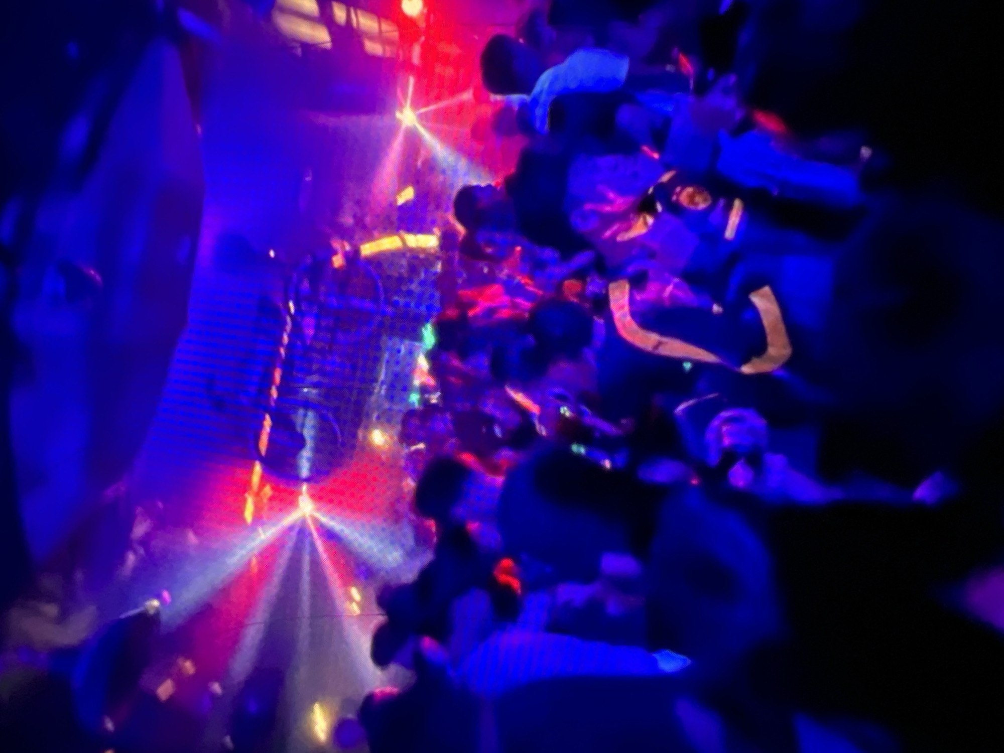 A crowded, dimly lit nightclub in Lagos, Nigeria, is depicted in the image. The atmosphere is vibrant, with blue and pink lights creating a dynamic and energetic ambiance. The club appears to be filled with people, many of whom are dancing and enjoying the music. The lighting from the stage casts beams of light across the room, illuminating the smoke-filled air and creating a visual spectacle. The crowd is diverse, with individuals of various ages and backgrounds, all united by their shared experience of the nightlife. The scene captures the essence of a lively night out in Lagos, a city known for its vibrant social scene and energetic nightlife.