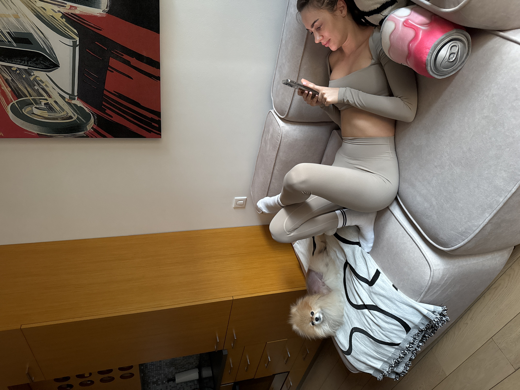 The image captures an indoor scene, likely in Warsaw, Poland, from a high-angle perspective looking down into a cozy living space. A young woman is the central subject, reclining comfortably on a light grey sofa. She is dressed in light-colored athletic wear, including a long-sleeved crop top and matching leggings, with white socks featuring black stripes. Her head is tilted, and she is engrossed in her smartphone, held in both hands.
Next to her head on the sofa is a distinctive decorative pillow shaped like a pink and white soda can with a metallic-looking top. At her feet, partially covered by a grey and white patterned blanket or throw, lies a fluffy, light-colored Pomeranian dog, looking directly towards the camera.
The background features a white wall with a large, vibrant piece of wall art depicting an abstract or stylized automotive scene in red, black, and white. Below the artwork, a wooden cabinet or storage unit with multiple drawers extends across the lower left of the frame. The floor appears to be wooden. The room is well-lit, suggesting daytime. The woman appears relaxed and focused on her phone, while the dog seems calm. No visible text is discernible in the image.