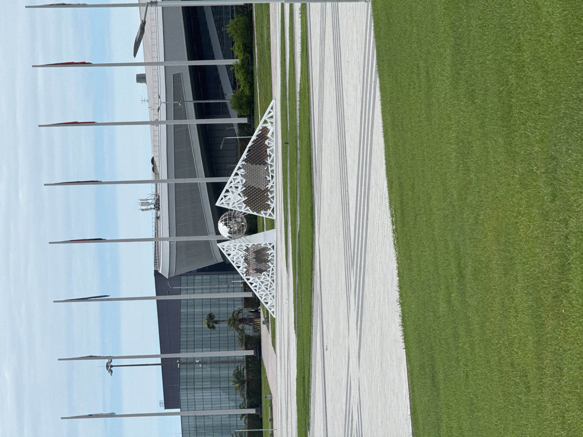 The image shows an outdoor scene in the city of Sirius, Russia. A modern building with a glass facade and a metallic roof is visible. In front of the building are tall flagpoles with flags. A white, triangular art installation with a globe on top stands alongside a paved pathway. The pathway is bordered by strips of green grass. The sky is partly cloudy. No people are clearly visible.