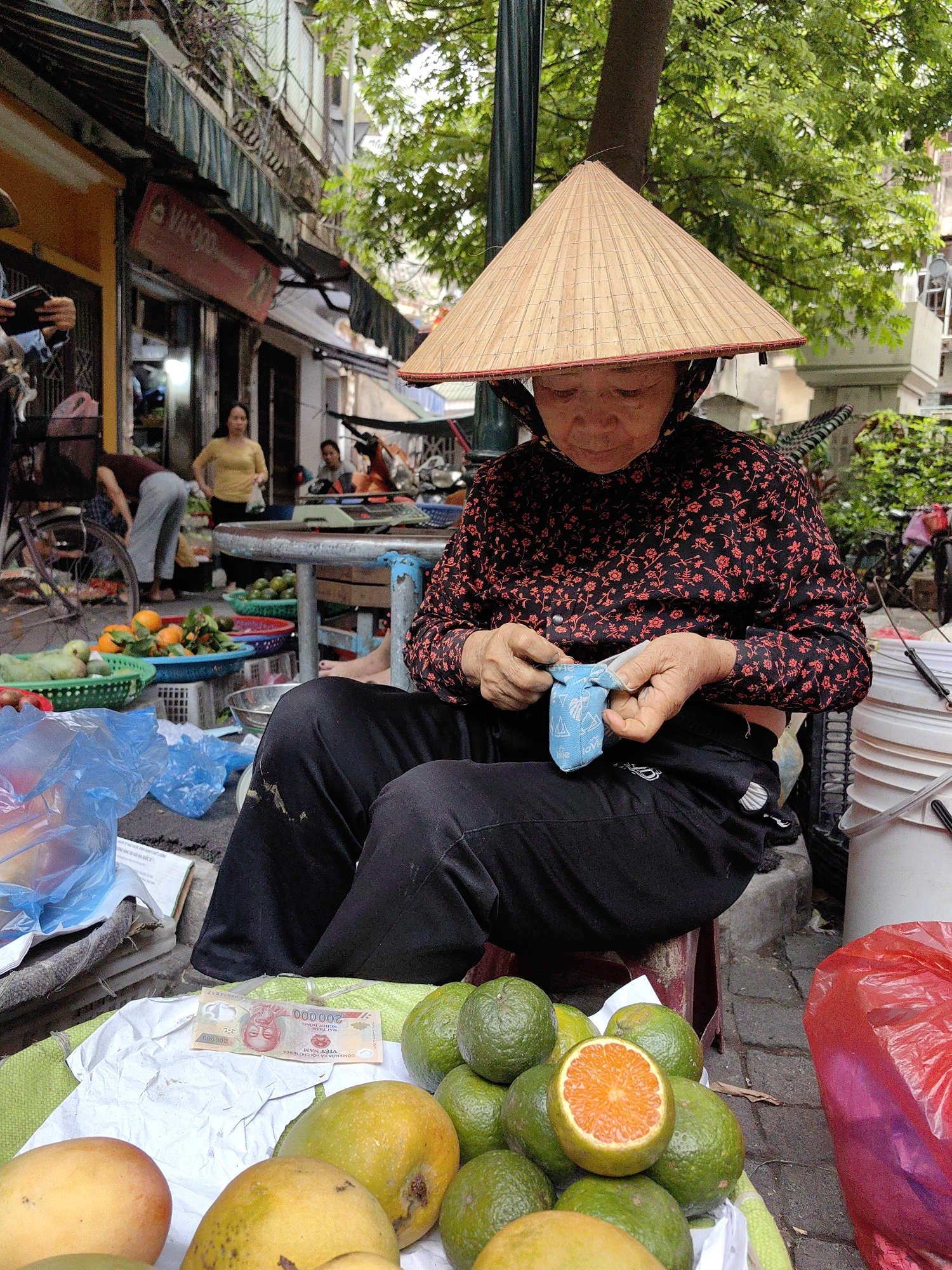 An elderly woman, likely a street vendor, is the central subject of this image, set in a bustling street market in Hanoi, Vietnam. She is seated on a low stool, wearing a traditional Vietnamese conical hat (nón lá) and a long-sleeved black shirt adorned with a red floral pattern, paired with black trousers. Her attention is focused on a small, light blue patterned pouch she holds in her hands.
In front of her, an assortment of fresh fruits is displayed for sale, including ripe yellow mangoes and numerous green oranges or limes, with one orange cut open to reveal its vibrant orange interior. A 200,000 Vietnamese Dong banknote is visible on a white cloth amongst the fruits, bearing the text "200.000", "CỘNG HÒA XÃ HỘI CHỦ NGHĨA VIỆT NAM", and "NGÂN HÀNG NHÀ NƯỚC VIỆT NAM". Blue plastic bags and other vendor items are also scattered around her.
The background shows a lively street scene under what appears to be an overcast or shaded daytime sky, with lush green trees overhead. Several other people are visible, including one person in a yellow top standing near a table and another leaning down towards baskets of fruit. Buildings with awnings line the street, contributing to the urban market atmosphere. A partial view of a bicycle and other market activities can be discerned. A red sign with "VAFOOD" is visible in the distance. The scene captures a slice of daily life and commerce in Hanoi.