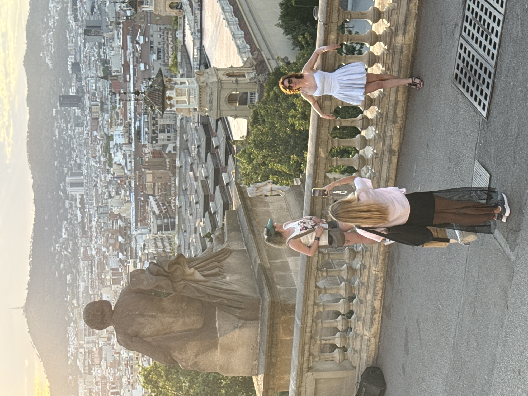 The image captures a scenic overlook in Barcelona, Spain, with the city sprawling below and a mountain in the background under a bright sky. In the foreground, a large stone statue stands to the left, facing away from the viewer.
Three women are present. One woman, wearing a white tank top, denim shorts, and a green visor, stands near the railing, facing away from the camera and looking out at the view. Another woman, dressed in a short white flared dress and sunglasses, leans against the railing, also looking away from the camera. The third woman, with blonde hair, is closer to the viewer and is taking a picture of the woman in the white dress with her phone. She wears a light pink shirt and a black skirt. A black bag is on the ground to the left, and another black bag is slung over the shoulder of the woman in the pink shirt.
The railing in the foreground is made of stone and features decorative balusters. Beyond the railing, the city's rooftops and buildings stretch out, with some taller structures and a distinctive tower visible. Lush green trees are scattered throughout the urban landscape. The ground is paved with asphalt.