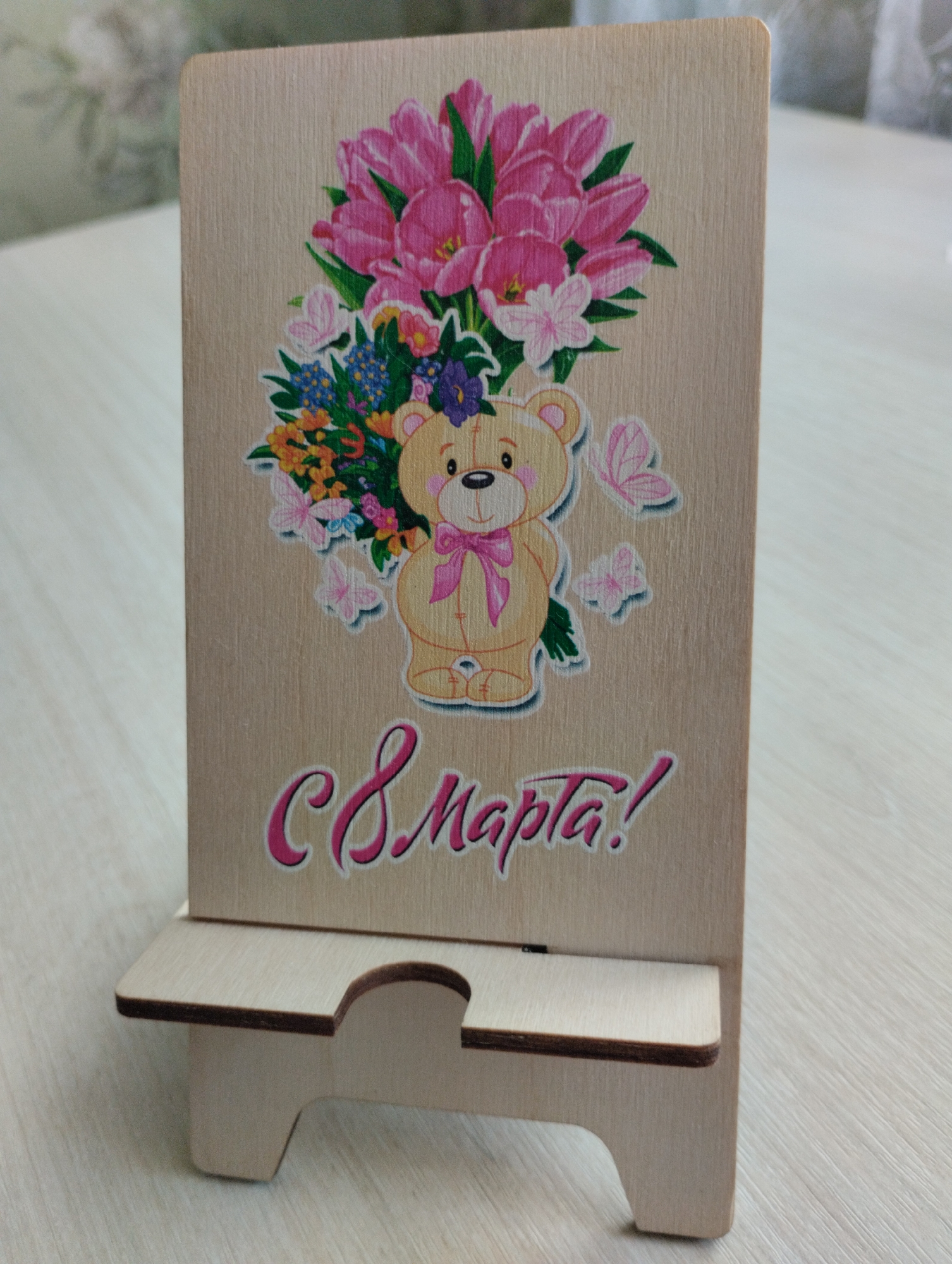 A close-up, eye-level shot shows a wooden stand for a mobile device, decorated with a graphic of a teddy bear holding a bouquet of pink tulips and smaller flowers. Pink butterflies are also part of the graphic, and text in a cursive font reads "8 Марта!" which translates to "March 8th!". The stand itself is made of light-colored wood with a cutout for a phone to rest. The background is blurred, suggesting an indoor setting with a light-colored surface, possibly a table. The image is sharp and well-lit, focusing on the details of the stand and its decoration. There are no people or animals present in the image. The location is not specified within the provided image or context.