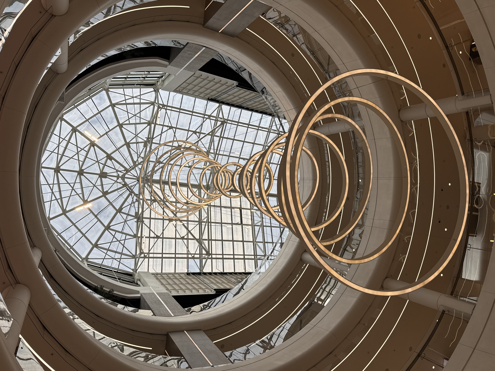 The image captures an interior view of a multi-level building, likely a shopping mall or atrium, in St. Petersburg, Russia. The perspective is looking upwards through a circular opening, revealing a complex architectural design. The building is constructed with concentric circular floors or levels, each finished with a beige or light gray material. These levels have recessed linear lighting elements that add a modern touch. A series of illuminated golden circular rings are suspended from the ceiling, creating a dynamic and artistic focal point. Above this sculptural element, the building features a large glazed skylight, supported by a metal framework in a grid pattern. This structure allows natural light to filter into the space. Two levels are visible with passageways.