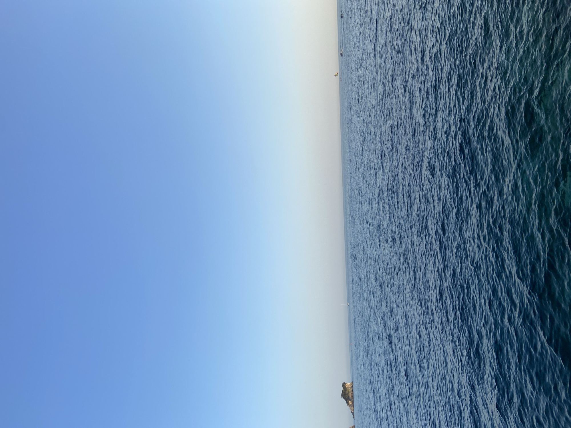 A wide shot captures a vast expanse of deep blue ocean under a clear, light blue sky. The water is slightly choppy, with gentle waves creating a textured surface. In the distance, a small, rocky island covered in greenery is visible on the left side of the frame. Further to the right, a parasailer is being pulled by a boat, with several other boats also present on the horizon. The overall scene is serene and depicts a typical day at sea.