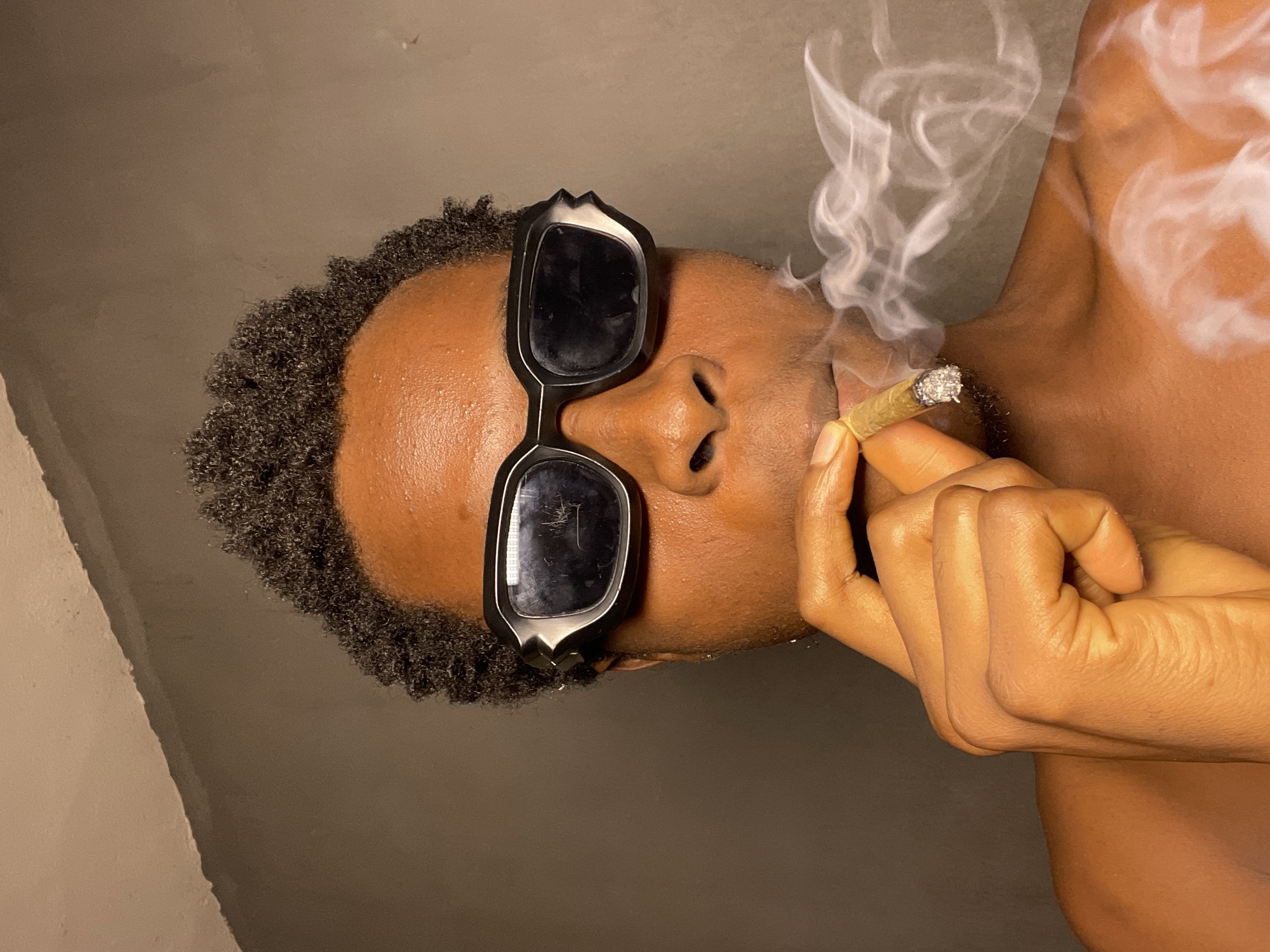 A close-up, waist-up, frontal shot shows a person with dark skin and short, curly hair wearing distinctive black and white sunglasses. The person is holding a lit cigarette, from which smoke is emanating and obscuring the lower half of their face and chest. The background is a plain, dark gray wall. No specific location in Nkpor, Nigeria is discernible from the image.