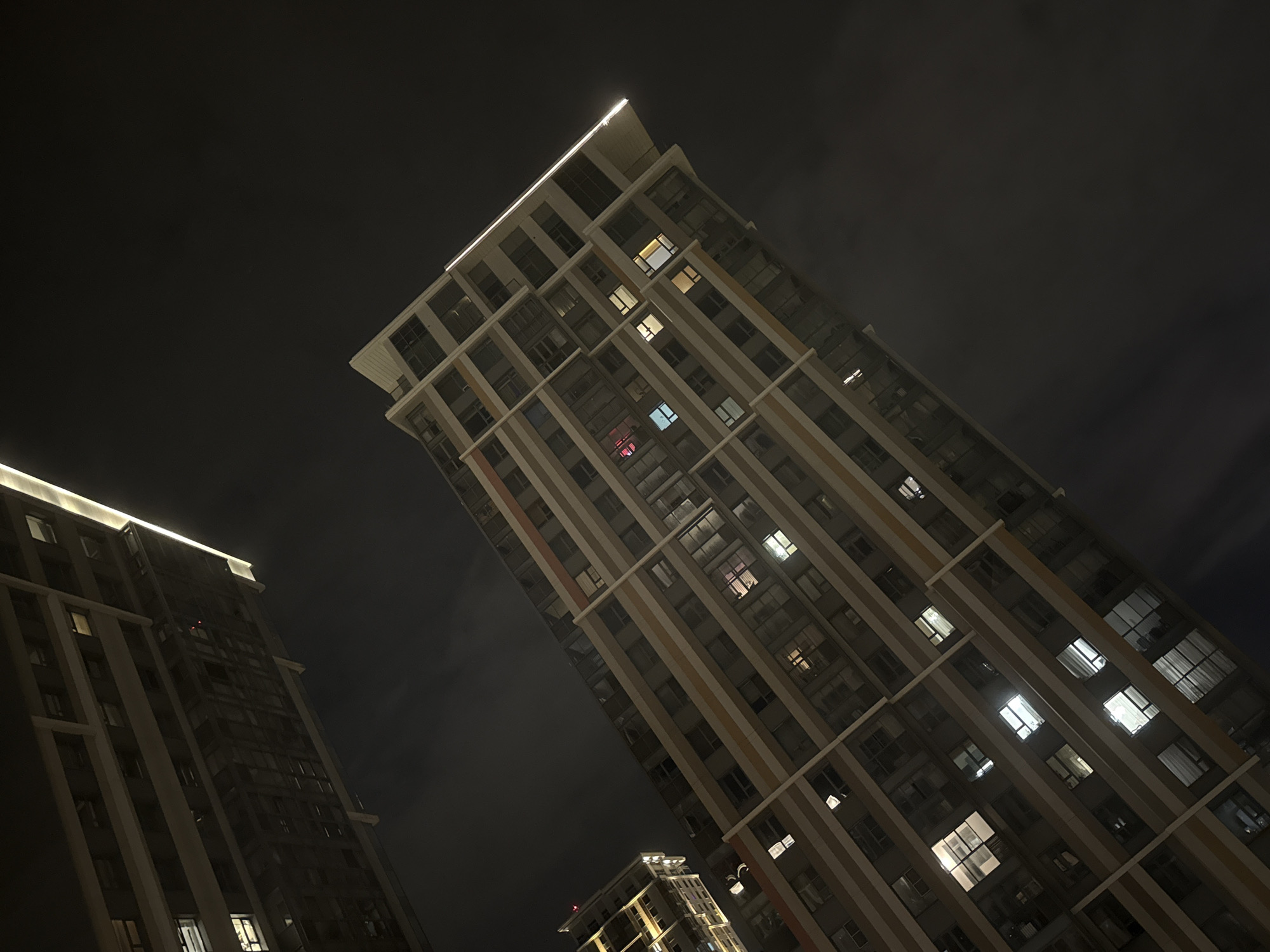 The image depicts a low-angle view of three modern high-rise buildings at night in Khimki, Russia. The buildings are lit, with some windows showing interior lights. The sky is dark, with indistinct clouds. The perspective is skewed, making the buildings appear to lean towards the right. The buildings have a light-colored facade with vertical stripes. Illumination is also present at the tops of the buildings. No people or other subjects are visible.