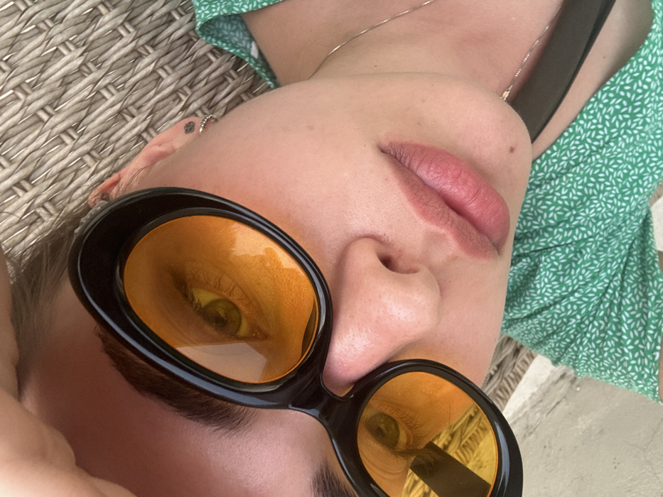 A close-up, high-angle portrait shot depicts a female subject from the chest up, positioned against a textured, woven background. The subject is wearing black-framed, oval sunglasses with bright yellow-orange tinted lenses through which her green or hazel eyes are visible. Her right hand is partially visible, resting on the upper left portion of her forehead. Her lips are a natural pinkish hue, slightly parted. A small, dark stud earring is visible on her right earlobe, and a small dark tattoo is present near the ear. She wears a green garment with a white, small-patterned floral or leaf design and a thin, silver-colored necklace chain. A small mole is visible on the right side of her chin. The background consists primarily of a woven, wicker-like surface, with a lighter, possibly paved or tiled surface visible in the lower left quadrant. The scene is located in Unknown, Unknown.