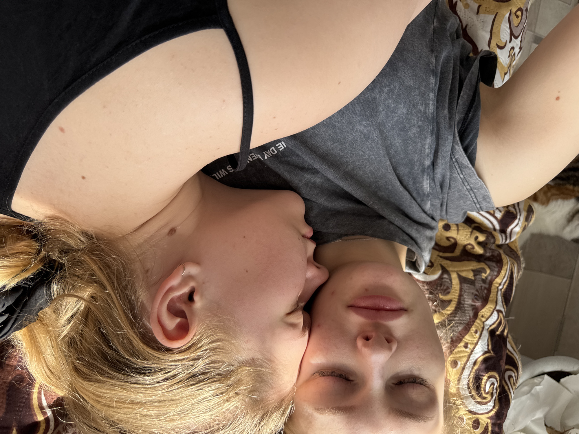 Two young women are lying head-to-head on a patterned surface, possibly a bed or blanket. One woman has blonde hair and is wearing a black tank top. The other woman has blonde hair and is wearing a gray t-shirt with text on the collar. The woman on the left is kissing the cheek of the woman on the right, who has her eyes closed. The location is unknown and cannot be determined as Бородулино, Russia from the image.
