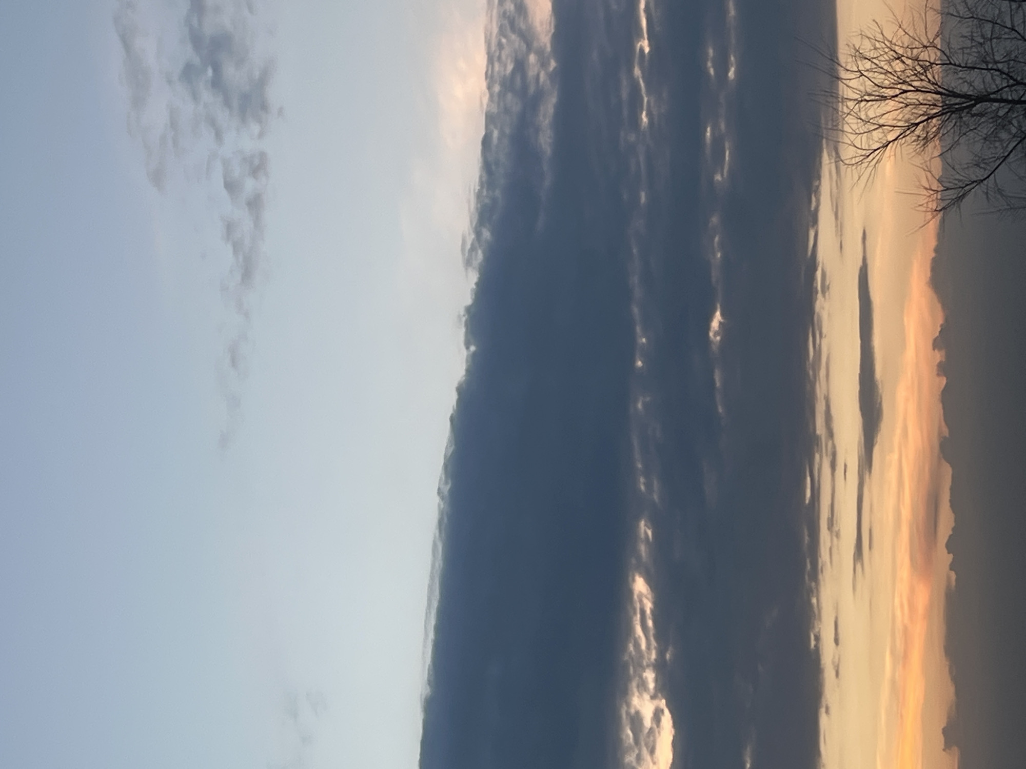The image shows a twilight sky with a mix of light blue and dark grey clouds. The sun appears to be setting, casting a golden glow on some of the clouds. Bare tree branches are visible in the upper right corner of the image. There is no explicit information about the location being Votkinsk, Russia, nor are there any people, actions, or objects specifically identifiable in the image that would allow for forensic or matter-of-fact descriptions pertaining to the city.