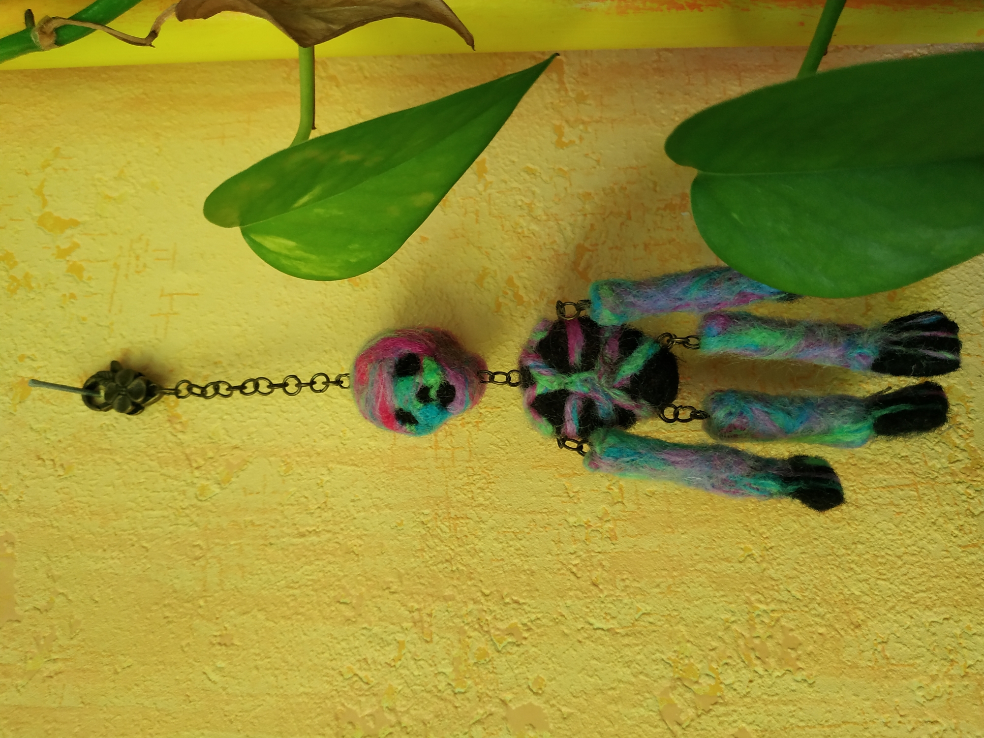 The image depicts a handmade felted wool doll with jointed limbs lying on a textured yellow surface. The doll has a round head with black spots, connected by a chain to a floral-shaped metal piece. The doll's body is also round, with four limbs attached at the sides. The limbs are yarn-covered and end in black felted extremities. Green houseplant leaves are visible in the upper portion of the image. The location is not determinable from the image and therefore the location in Dortmund, Germany can't be defined.