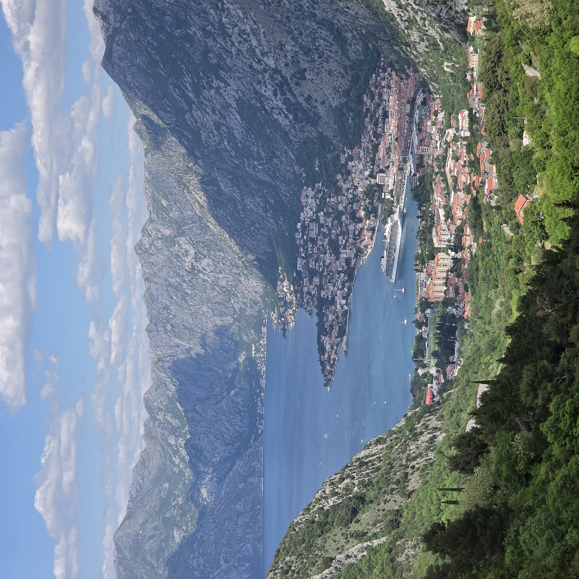 The image presents an elevated view of Kotor Bay, Montenegro, likely captured from a vantage point in the mountains. The deep blue waters of the bay stretch between steep, rugged mountainsides, with one mountain range prominently featured on the left, partly obscured by clouds.
Buildings with terracotta roofs cascade down the slopes on the right, transitioning into lush green vegetation. A large cruise ship is docked in the bay near the town, suggesting tourism activity. Small boats dot the water's surface. The sky above shows patches of blue interspersed with white clouds.