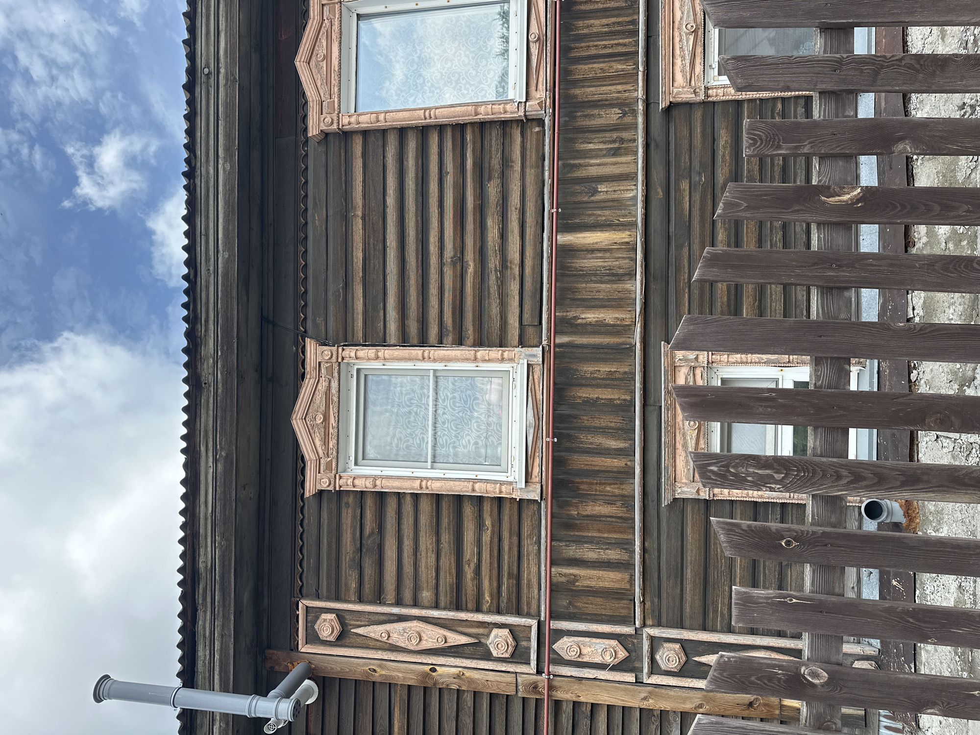 This is a medium shot, low-angle, exterior photograph of a wooden house with decorative window frames and a corrugated metal roof. The house has horizontally oriented wooden siding. There are two windows visible on the upper story, each with ornate pink trim. A vertical wooden decorative element with carved circular and diamond motifs is attached to the left side of the house. A grey pipe extends from the left side of the roof. A dark brown wooden fence made of vertical planks is in the foreground. The sky above is cloudy with patches of blue. The scene appears to be in Nogornyi, Russia. There are no people or animals in the image.