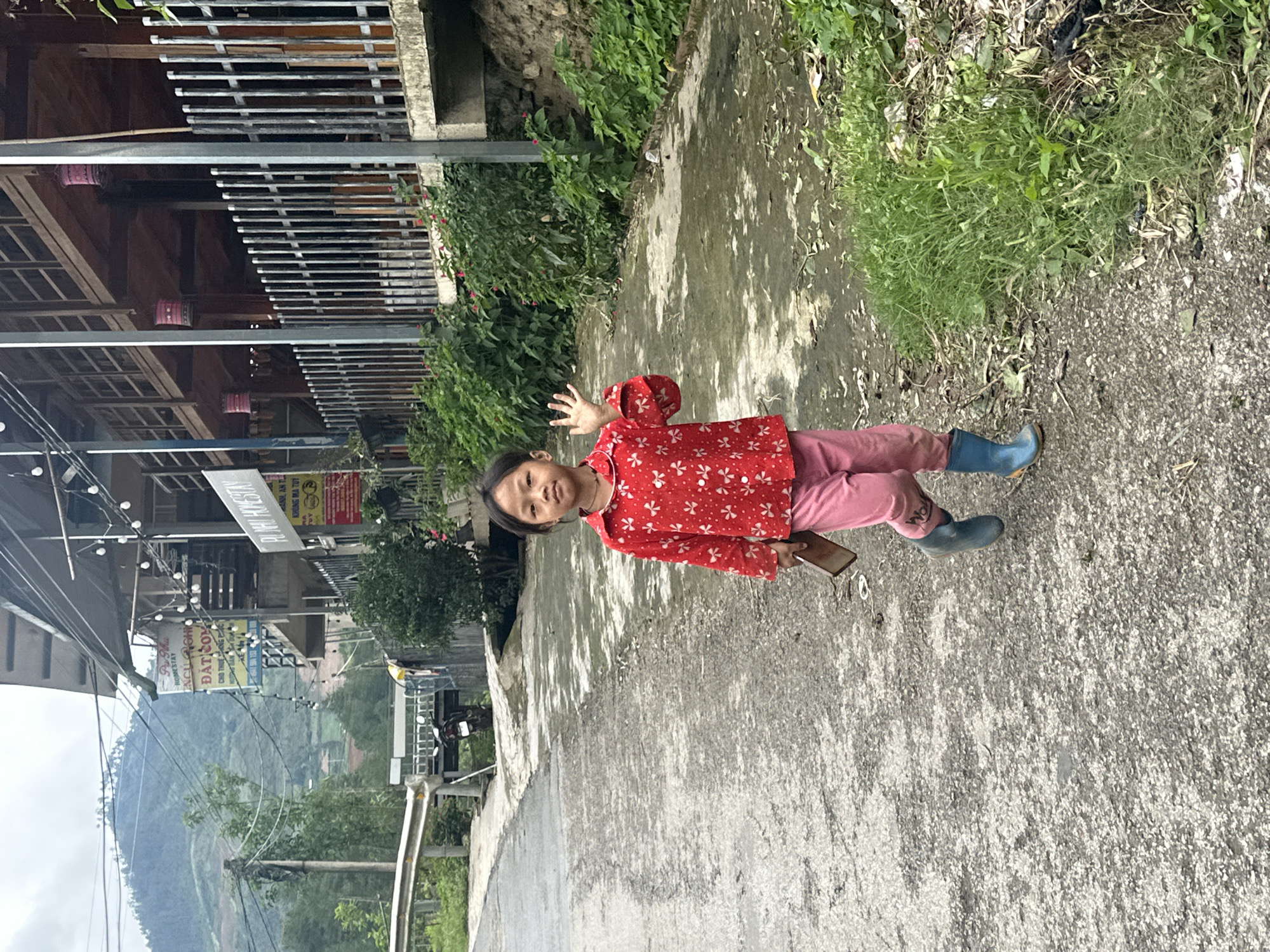 A young Asian child, approximately 4-7 years old, walks towards the viewer on a damp, grey asphalt road in Mù Cang Chải, Vietnam. The child wears a red long-sleeved shirt with white floral patterns, pink trousers, and blue and orange rubber boots. Their dark hair is cut short, and they wear a thin dark necklace. In their left hand, they hold a dark brown rectangular object, possibly a phone or wallet, while their right hand is raised in a wave. Their facial expression is neutral, eyes directed at the camera.
To the child's left, the road curves gently upwards, bordered by a metal guardrail and a concrete culvert structure under which a black motorcycle is partially parked. Further left, a utility pole supports multiple overhead power lines, set against a backdrop of steep, tree-covered mountains, some revealing terraced agricultural plots.
To the child's right, the road meets a strip of green grass and dense vegetation, including bushes with small pink flowers, leading to a dirt embankment. Above this embankment stand multi-story buildings, characteristic of homestays, featuring wooden upper levels with balconies and vertical metal railings. Several Vietnamese signs are visible on the buildings; one clearly reads "PÙ NHÌ HOMESTAY," and another includes "ĐẶT CƠM" (Order Rice/Food). Decorative string lights and traditional pink cylindrical lanterns hang from the building exteriors. The sky is overcast, casting a soft, diffuse light over the scene.