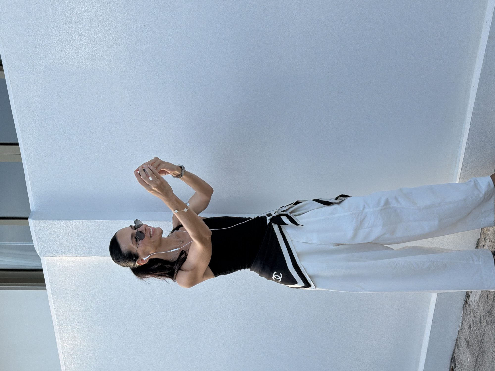 A woman with dark hair pulled back in a ponytail stands in front of a white textured wall in Ko Samui District, Thailand. She is wearing a black tank top, wide-leg white pants, and a black and white patterned scarf tied around her waist, featuring a Chanel logo. She has on sunglasses and white earbuds. The woman is holding up her phone with both hands, appearing to take a selfie or record a video. She is smiling and looking at her phone. The background is a plain white wall, with a portion of a building's window visible at the top left. The ground is a grey concrete surface.