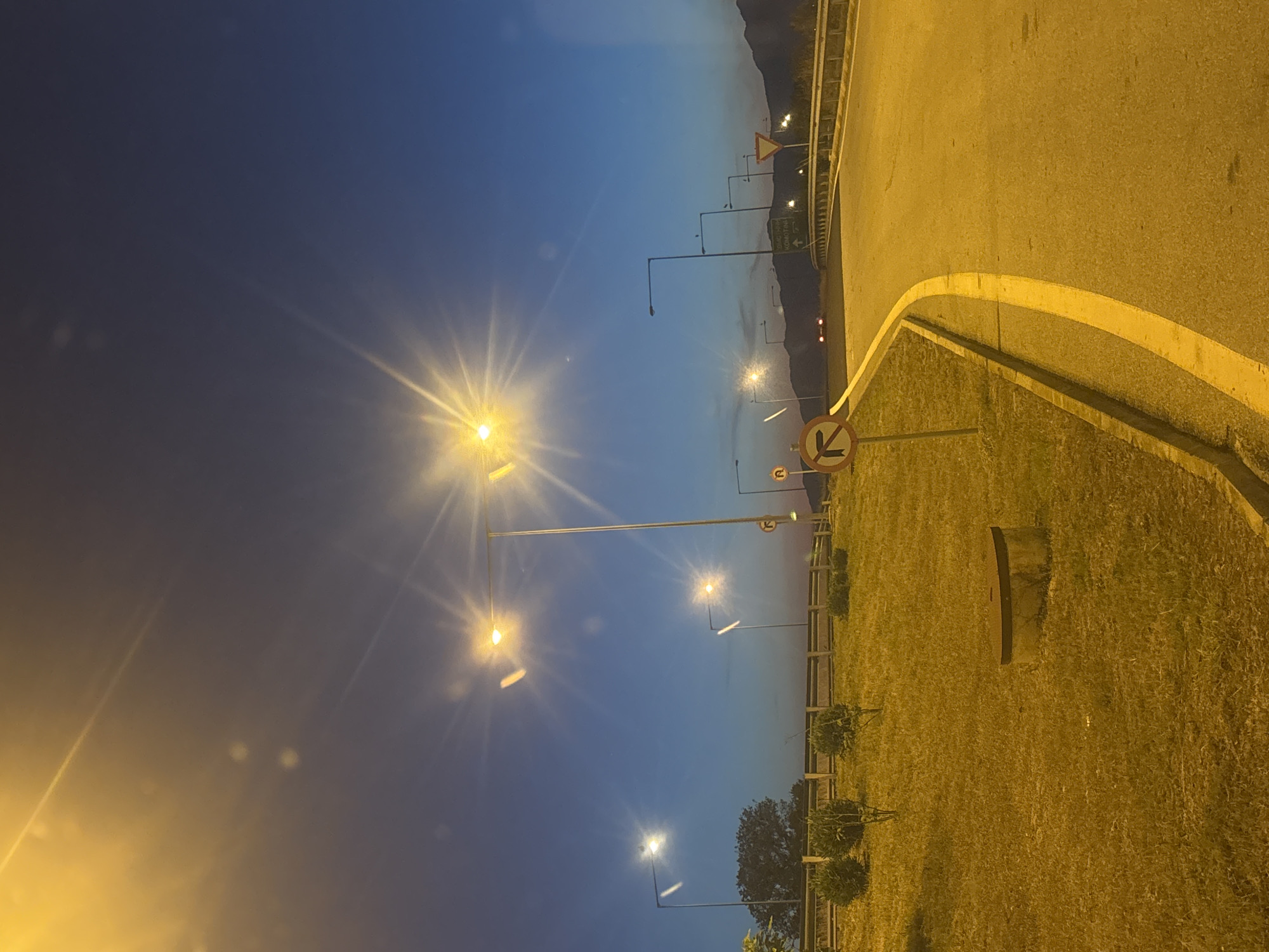 The image depicts a road intersection or highway exit at dusk or dawn, illuminated by streetlights. The sky is a deep blue fading to a lighter hue near the horizon, with bright starburst effects from the streetlights. The foreground features a grassy embankment with some low bushes. A circular concrete structure with a metal lid is visible on the grass. A paved road curves from the right of the frame towards the background. Several streetlights are positioned along the road and on the embankment. Road signs are visible, including a circular sign with a red border and a black arrow indicating a left turn restriction, and a triangular yellow sign with a black border. In the distance, mountains are silhouetted against the sky, and the headlights of a car are visible on the road. The scene appears to be in or near Venna, Greece, as indicated by a sign with Greek text that appears to say "KOMOTINI" with an arrow pointing forward. There are no people visible in the image.