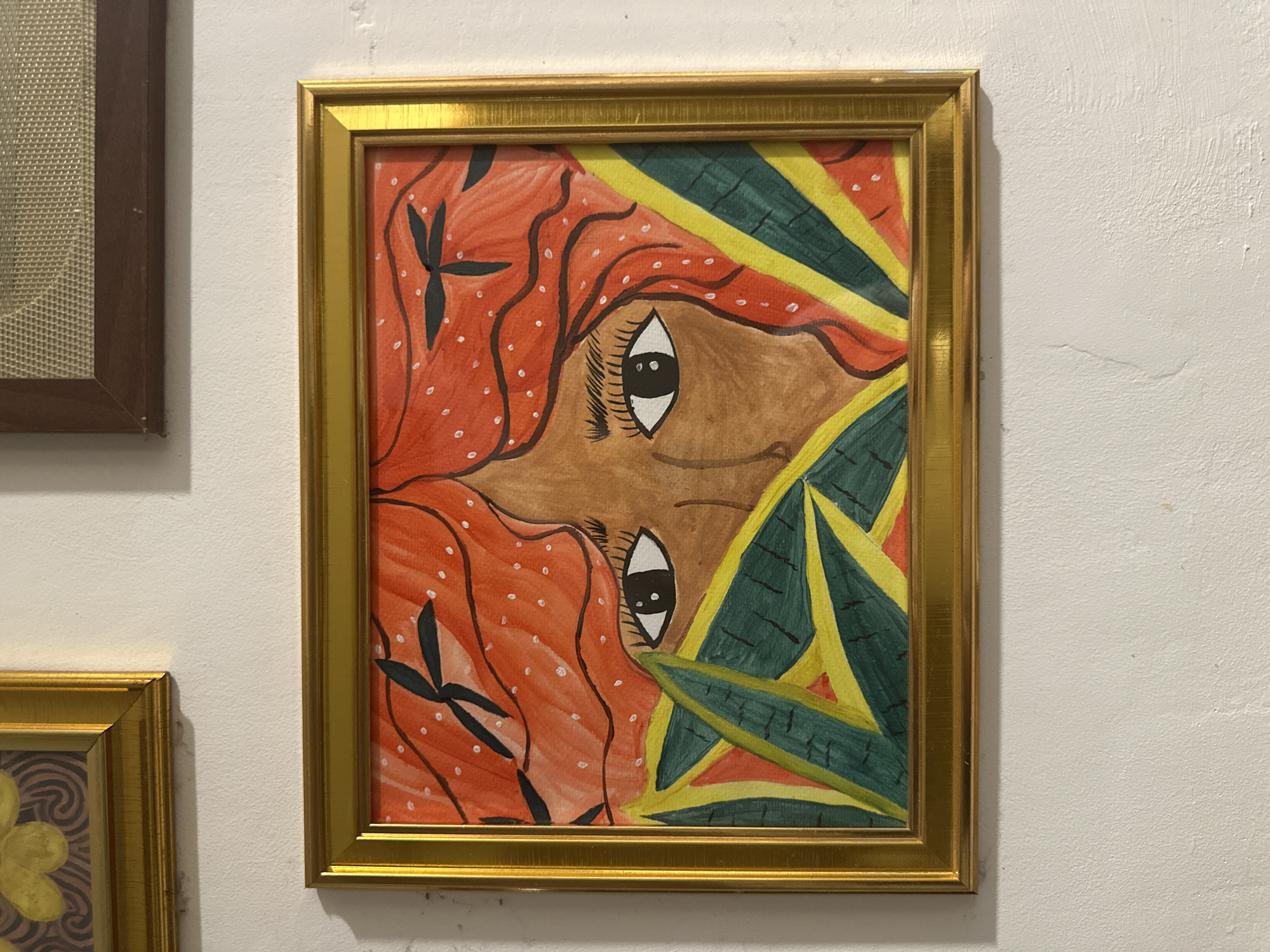 A vibrant, stylized painting in a golden frame is the central subject, hung on a white, textured wall. The artwork depicts a partial view of a face or head, characterized by brown skin and prominent, wide-open eyes with white sclera and black pupils. The hair is a striking orange, flowing, and adorned with numerous small white dots and intricate black patterns that resemble floral or leaf motifs. Surrounding the figure are bold, geometric patterns in shades of green, yellow, and hints of red, forming abstract or leafy designs.
The golden frame is sleek and reflective, encasing the colorful artwork. To the immediate left of the main painting, a portion of another framed artwork with a dark brown frame and a light, mesh-like or woven texture is visible. Below the central piece, the corner of a third artwork, also in a golden frame, can be seen, displaying a yellow, swirling floral pattern against a darker background.
The wall is painted white with a slightly uneven, textured finish, suggesting plaster or a similar material. The scene captures an indoor art display, possibly in a gallery or exhibition space, set in Davao City, Philippines. There is no visible text in the image.