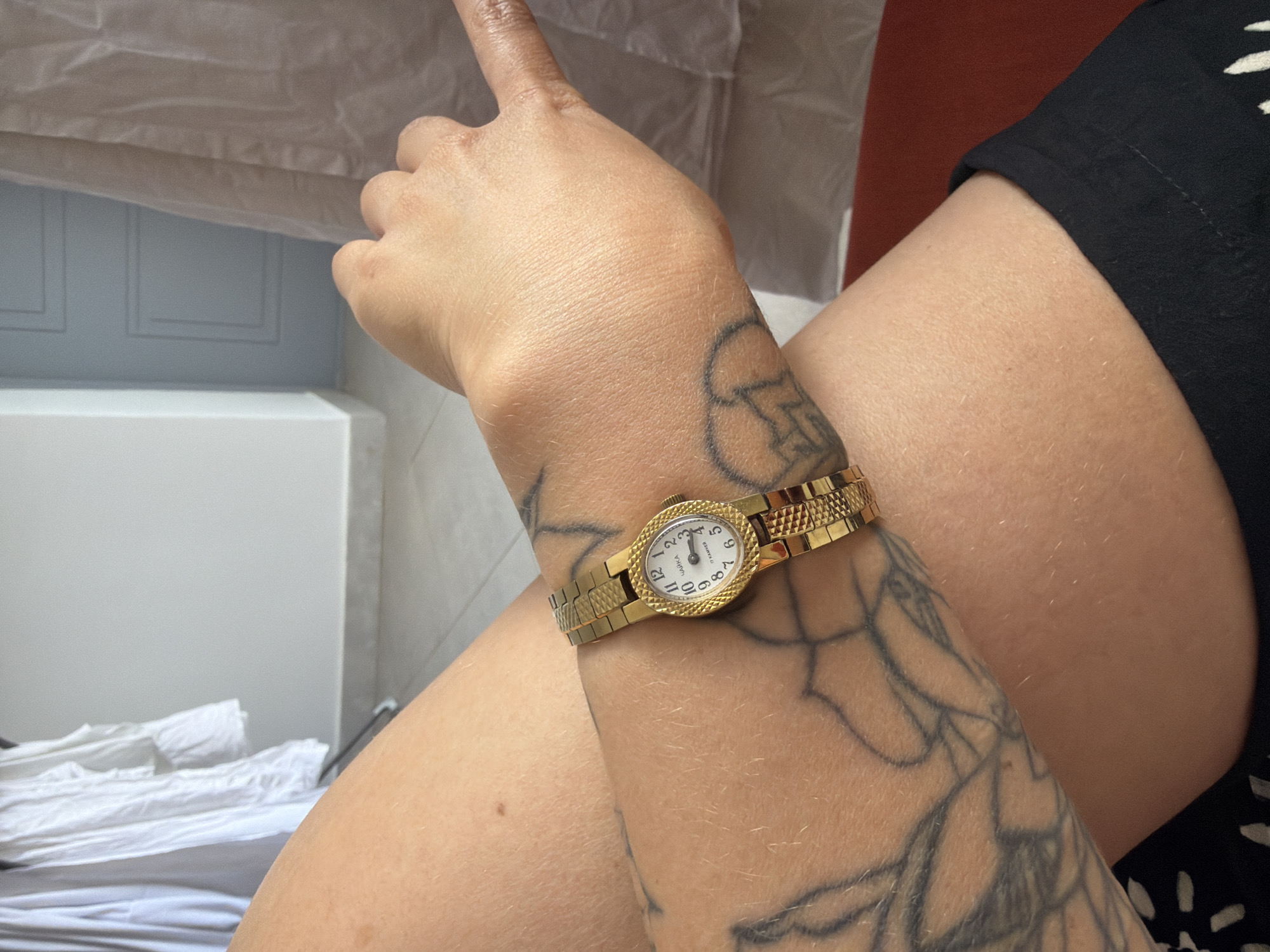 The image shows a close-up of a person's left forearm and wrist, adorned with a gold-colored wristwatch and tattoos. The watch, with a white face and Roman numerals, appears to be a vintage piece. The background is blurred, but hints of domestic elements like white curtains and a light-colored door suggest an indoor setting. There is no identifiable information to place the image in Sarapul, Russia.
