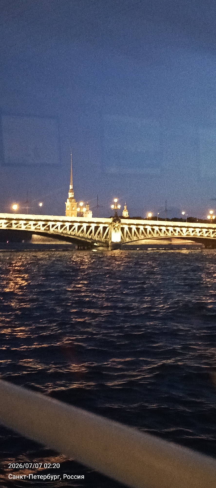 The image shows a nighttime view of a bridge and a spire in Saint Petersburg, Russia. The bridge, likely the Trinity Bridge, spans across a dark body of water, with its intricate metalwork illuminated by streetlights. In the background, a tall, slender spire, characteristic of the Peter and Paul Fortress, rises against the deep blue night sky. The water reflects the golden lights of the bridge and the sky, creating shimmering patterns. The lower right corner of the image shows a blurred foreground element, possibly part of a boat or viewing platform. Text overlays in the bottom left corner indicate the date and time as "2026/07/07 02:20" and the location as "Санкт-Петербург, Россия" (Saint Petersburg, Russia). There are no people visible in the frame. The scene captures a serene, illuminated cityscape at night.