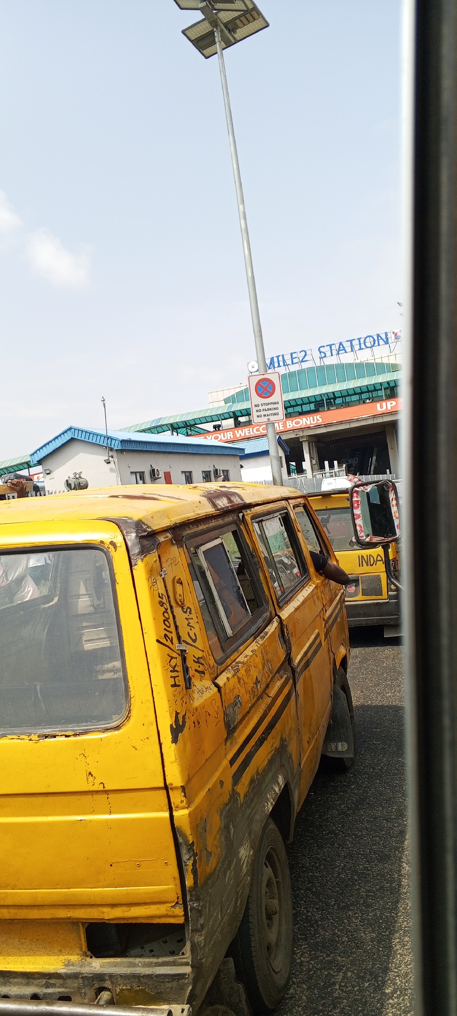 An eye-level, medium shot captures a yellow minibus in Lagos, Nigeria. The minibus, worn and weathered with visible rust and paint damage, occupies the foreground. Its windows are tinted, and part of its interior is visible, showing a passenger or driver. Writing, possibly a license plate or route information, is scrawled on the side of the vehicle. Another yellow vehicle, partially visible and identified by the word "IND...", is behind it. In the background, the "MILE 2 STATION" sign is prominently displayed on a modern-looking building with a turquoise roof and overhang. A lamppost with solar panels stands tall near the sign. A red and white circular traffic sign with a diagonal line through a red "X" and text below indicating "NO STOPPING, NO PARKING, NO WAITING" is also visible. The scene appears to be a street or road leading to or from the station, under a clear sky with scattered clouds. The overall impression is that of a bustling urban environment in a developing city.
