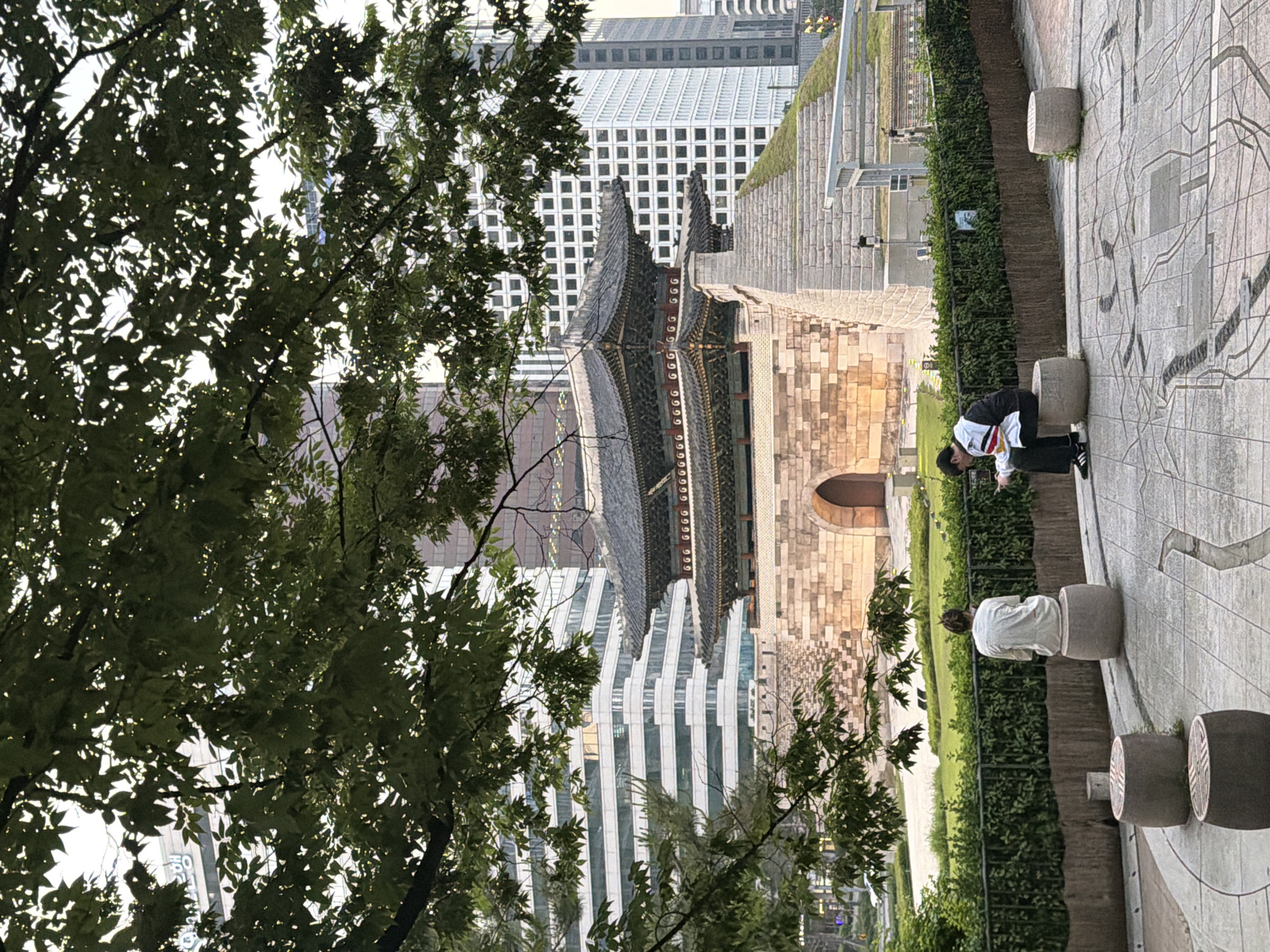 A rotated, medium shot shows two people located near an ancient stone-bricked structure, trees, and modern buildings, suggesting a blend of historical and contemporary elements. It seems that the shot was taken from a higher vantage point. In the foreground, a tree with dense green foliage occupies the left side of the frame.
Behind the tree, the focus shifts to an old, multi-tiered structure with distinctive East Asian architectural details, including ornate eaves and a tiled roof. This structure contrasts with the modern buildings in the background, one of which features a grid-like facade.
On the right side of the image, two people are present. One person, wearing a black and white jacket and black pants, is bending over, appearing to be looking at something on the ground. Another person, clad in a white shirt, is seated on a circular stone seat.
The ground is paved with a map design. The entire scene is bathed in daylight, indicating an outdoor setting. The scene is likely to be in Seoul, South Korea, because of the style of the pagoda and modern buildings in the area.
